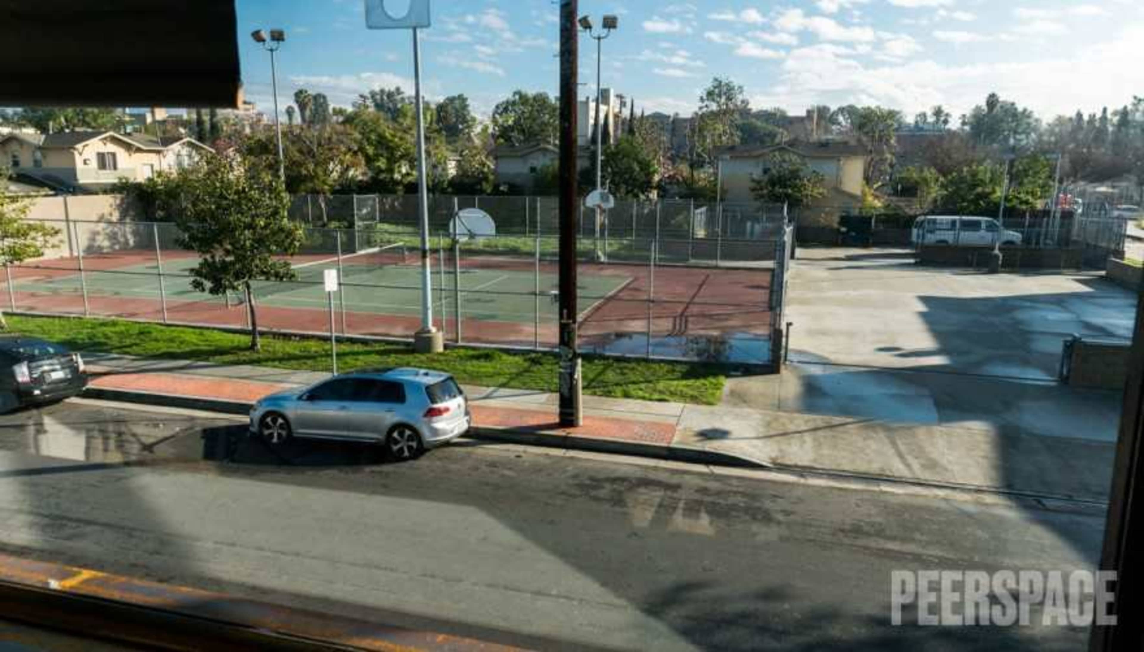The image shows a view of an outdoor tennis court area alongside a street with parked cars.
