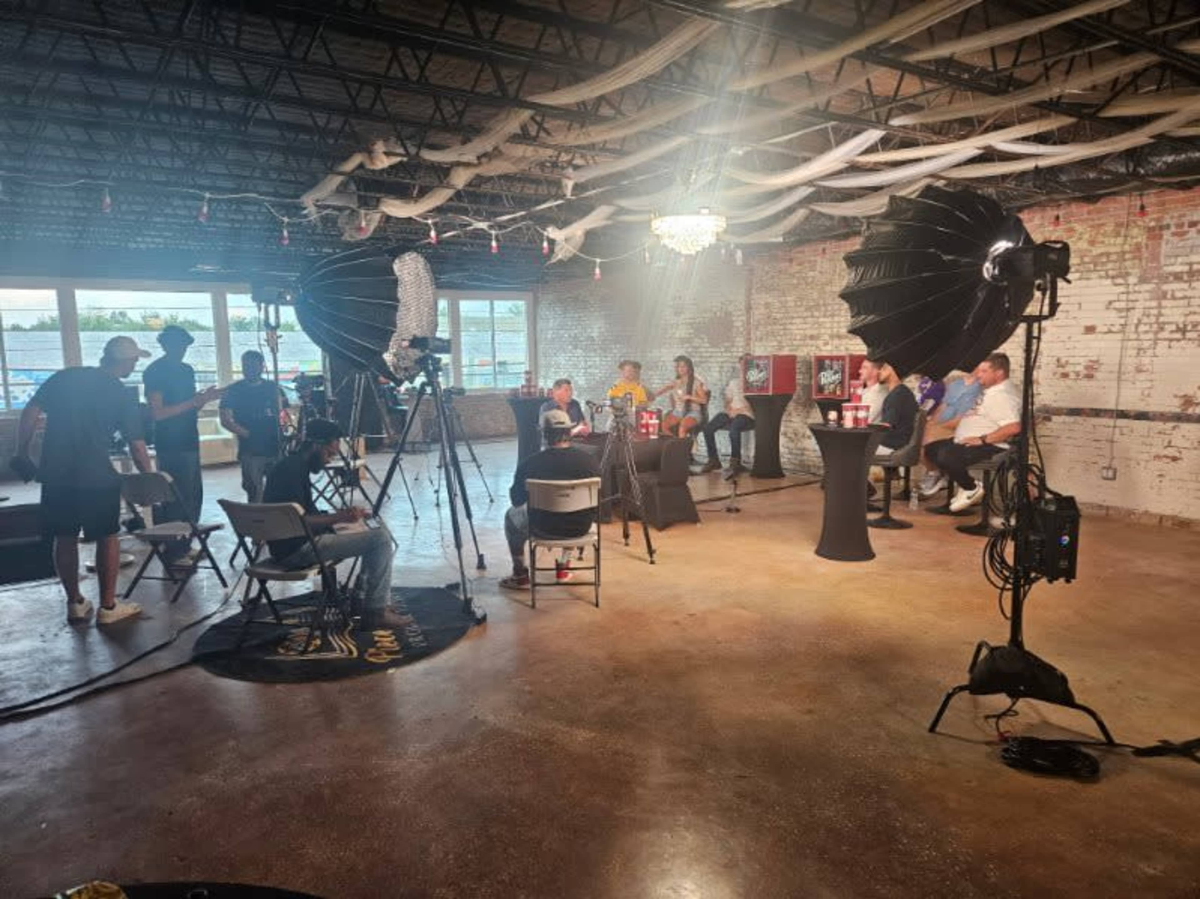 A group of people is seated around a table in a spacious, industrial-style room equipped with lighting and cameras for a filming setup.