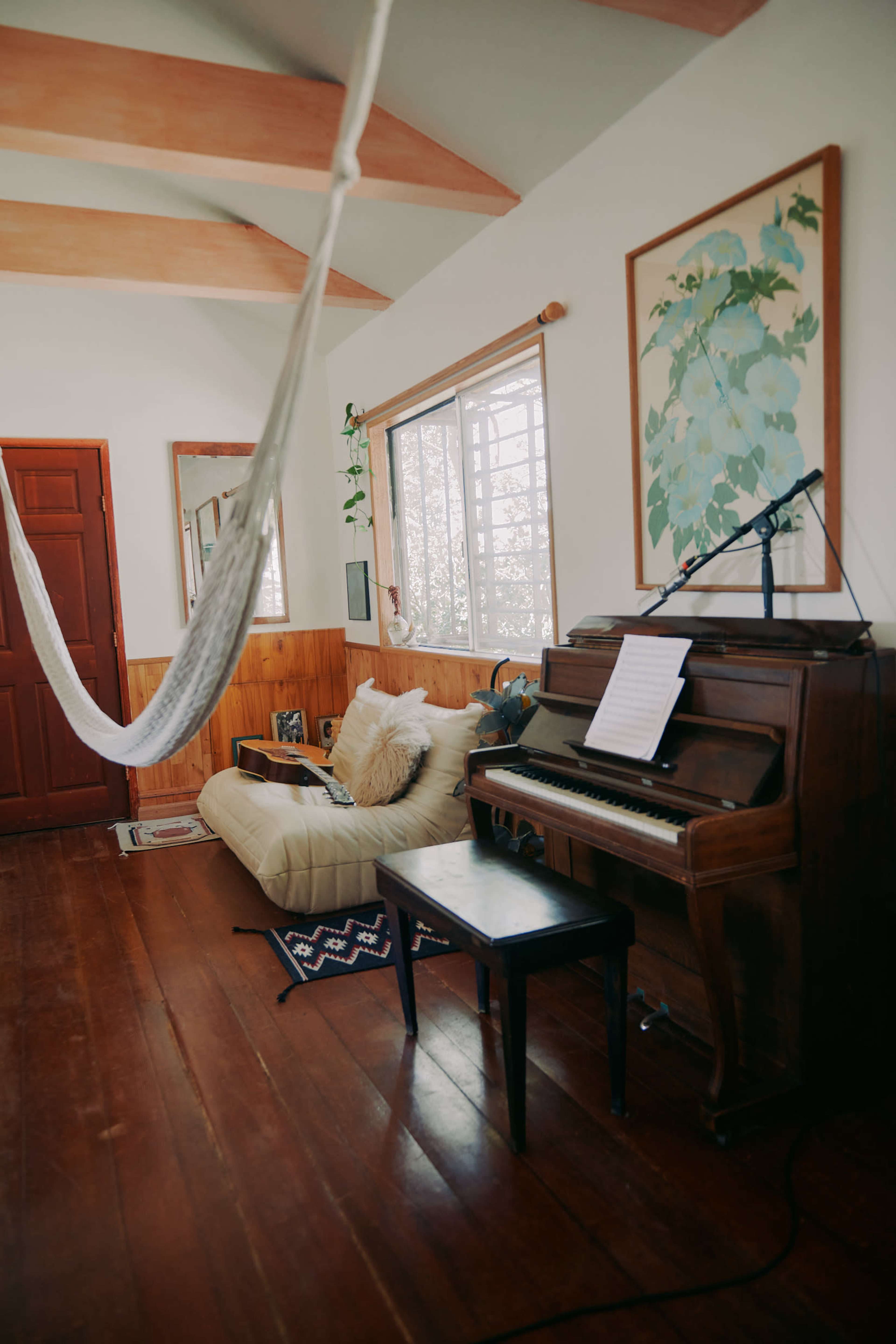 The image shows a cozy living room with a piano, a small table, a hammock, and large windows letting in natural light.