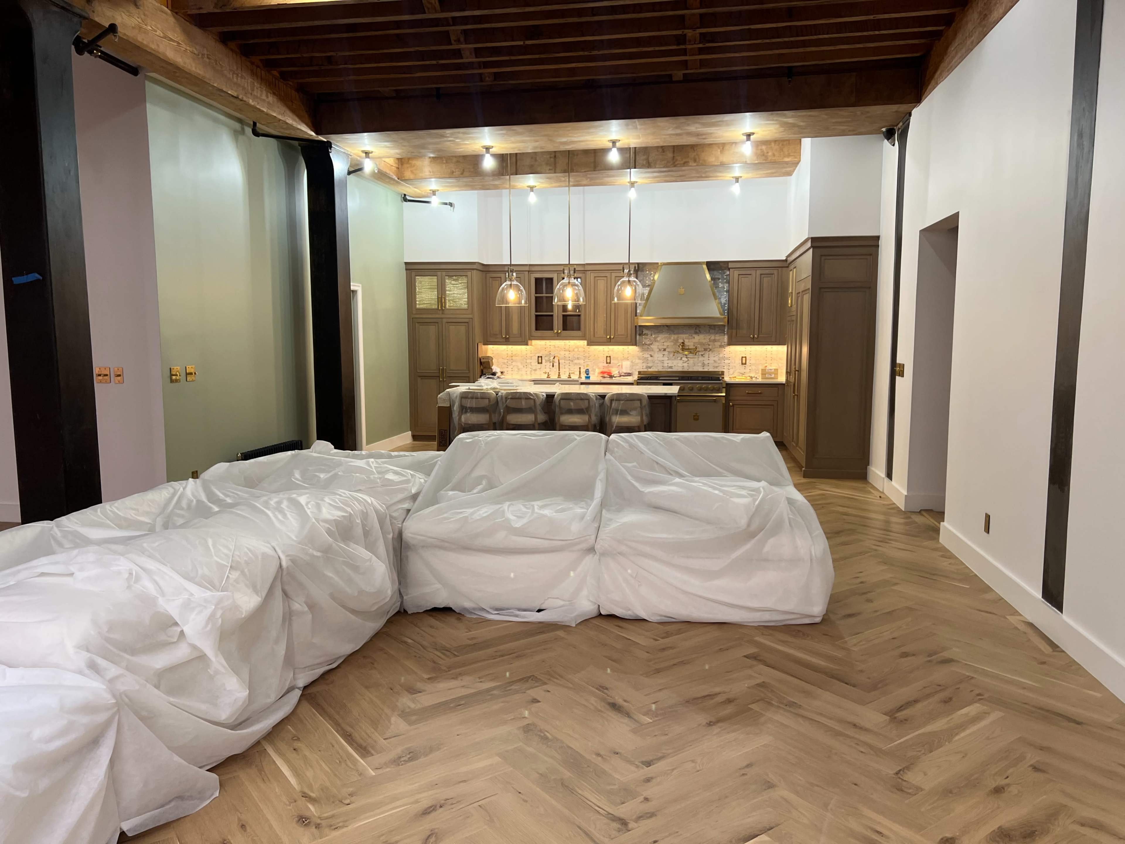 The image shows a partially furnished room with covered furniture and a kitchen in the background featuring wooden cabinets and countertop lighting.
