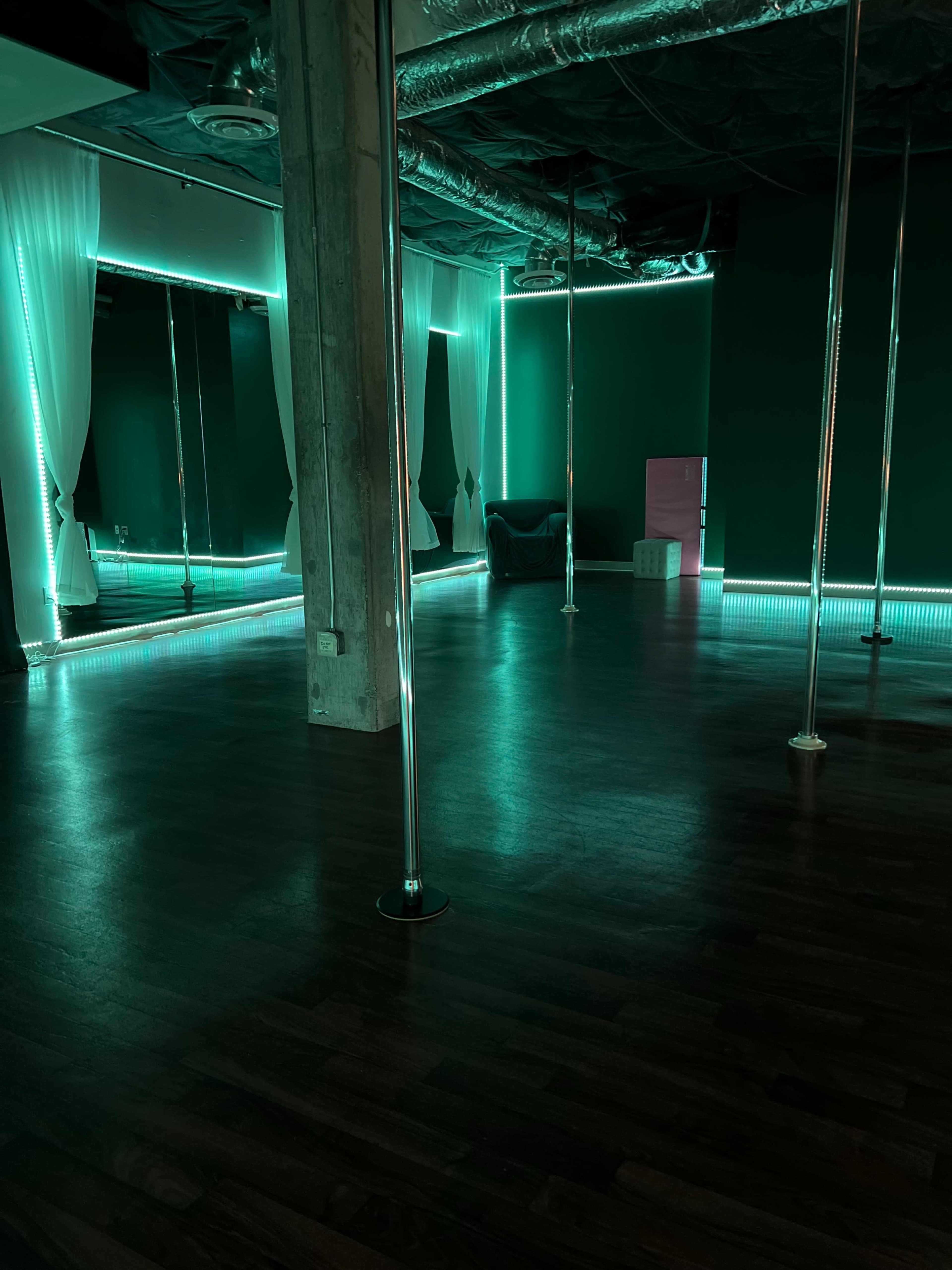 The image shows a dimly lit studio featuring several mirrored walls and chrome poles, illuminated by teal LED lights.
