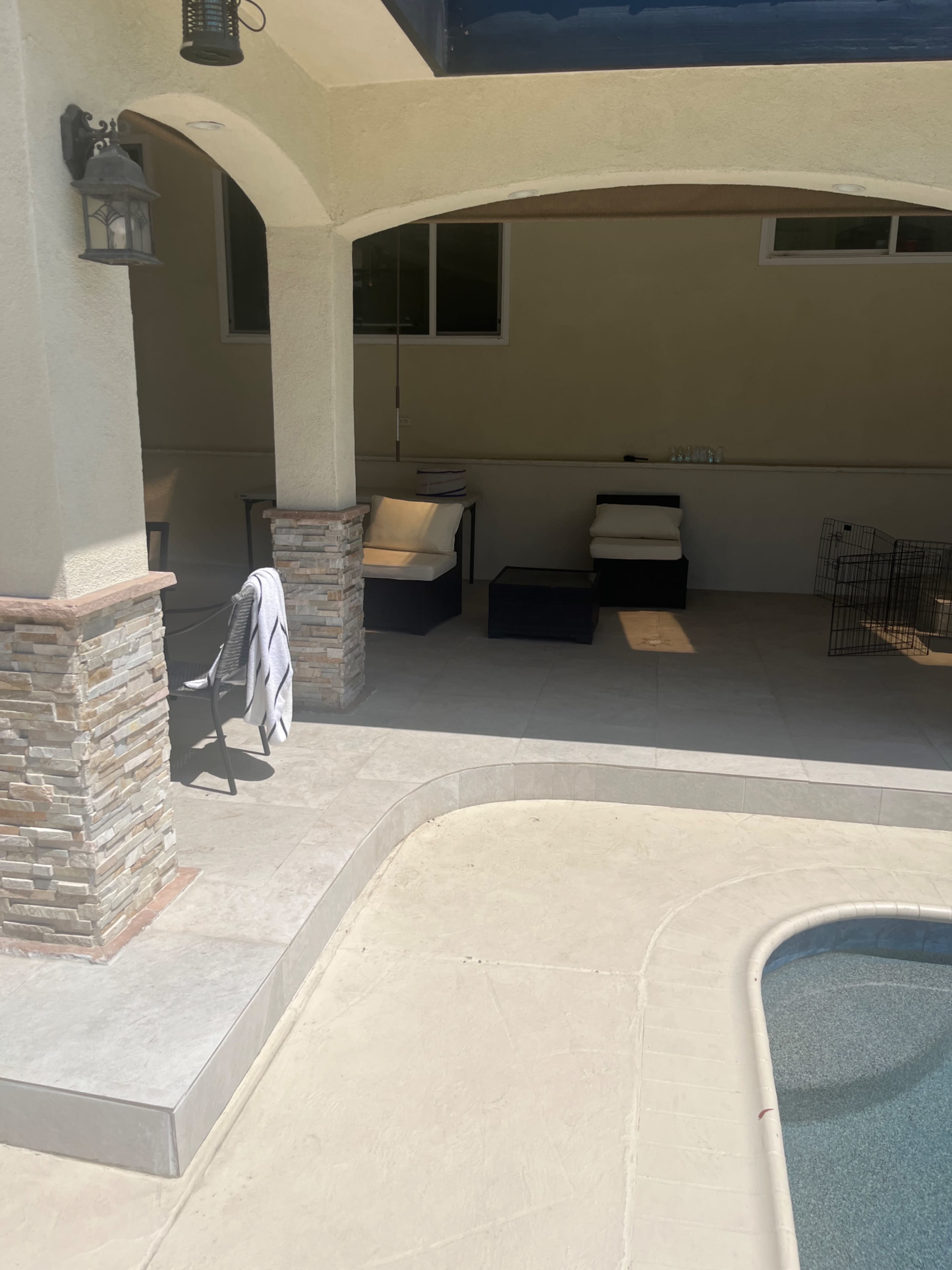 The image shows a shaded outdoor area with stone pillars, a tiled floor, and two seating areas near a swimming pool.