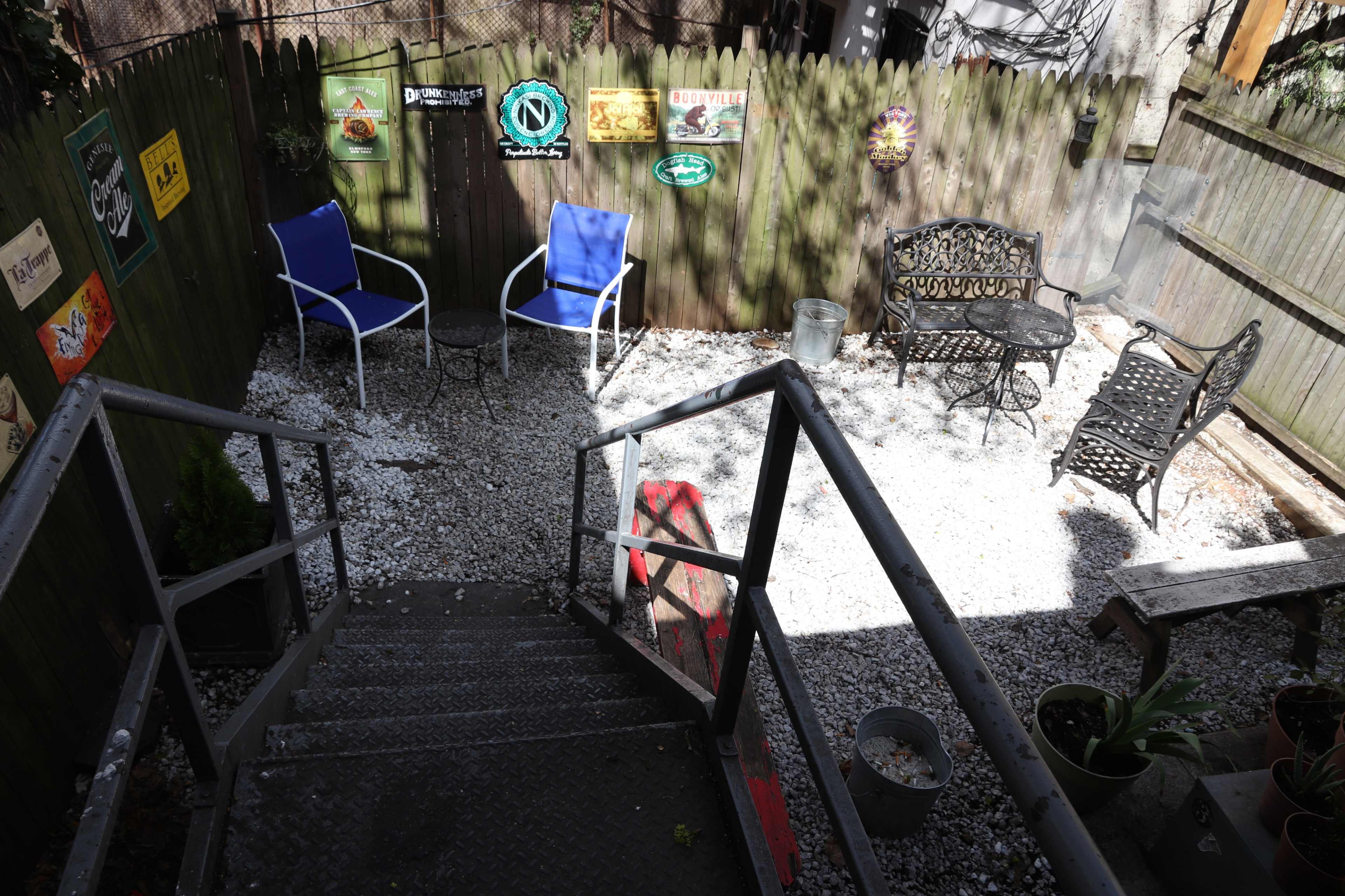 A gravel patio area with two blue chairs and a wrought iron table set against a wooden fence adorned with various signs.