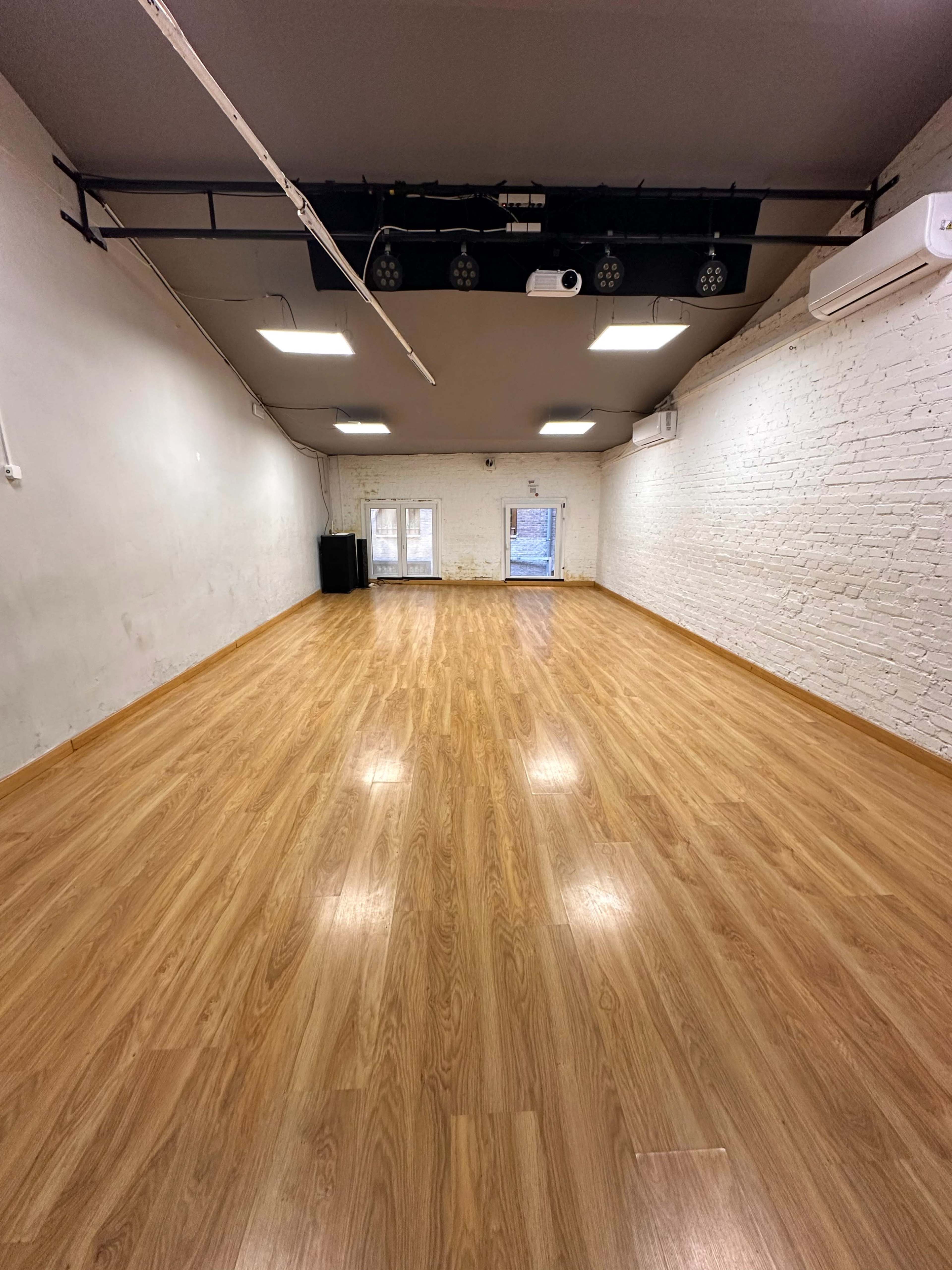 The image shows a large, empty room with wooden flooring, white brick walls, and overhead lighting, featuring two doors at the far end.