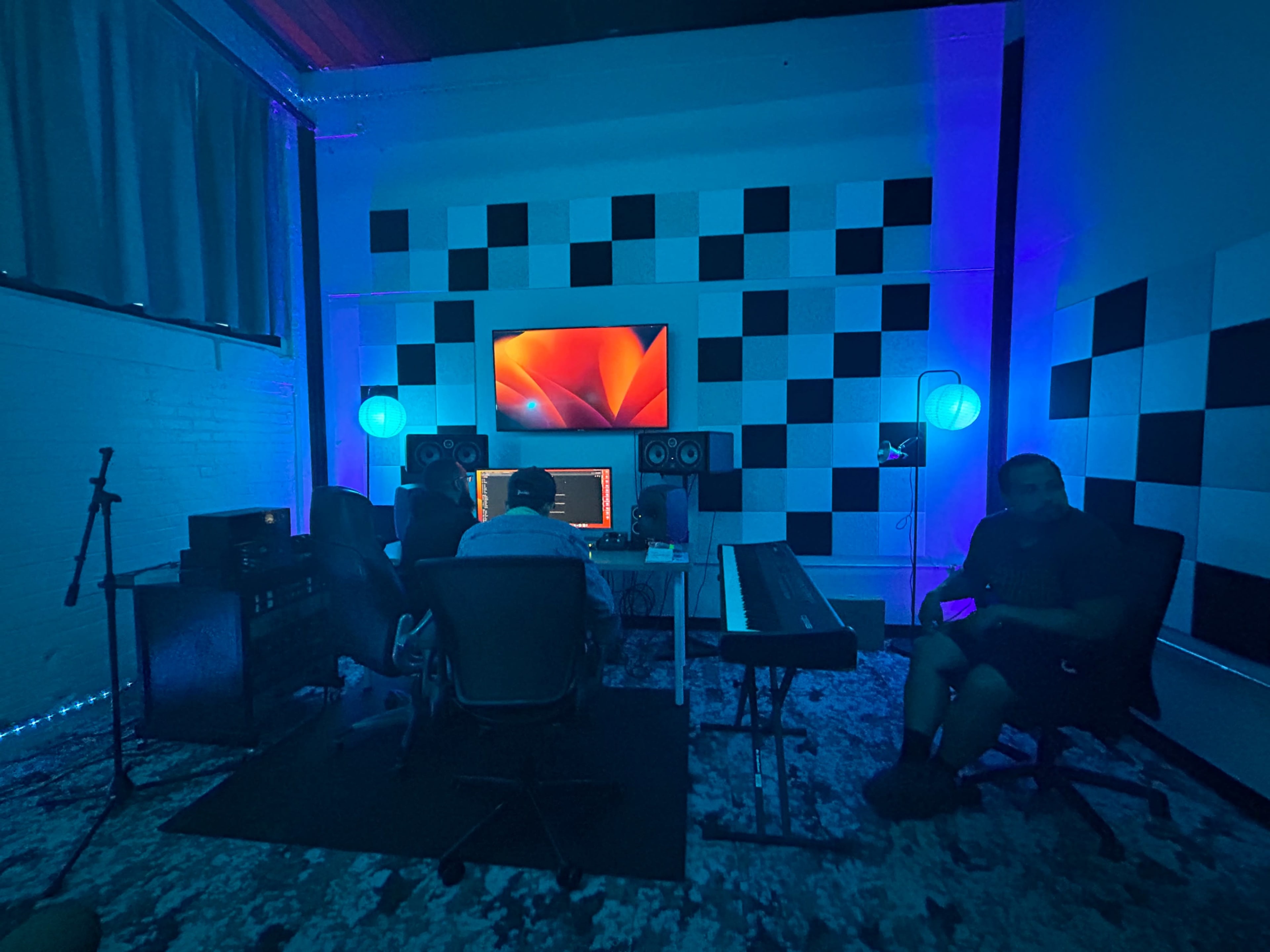 The image shows a modern music production studio with a brightly lit workspace featuring a computer, keyboard, and multiple speakers against a checkered wall.