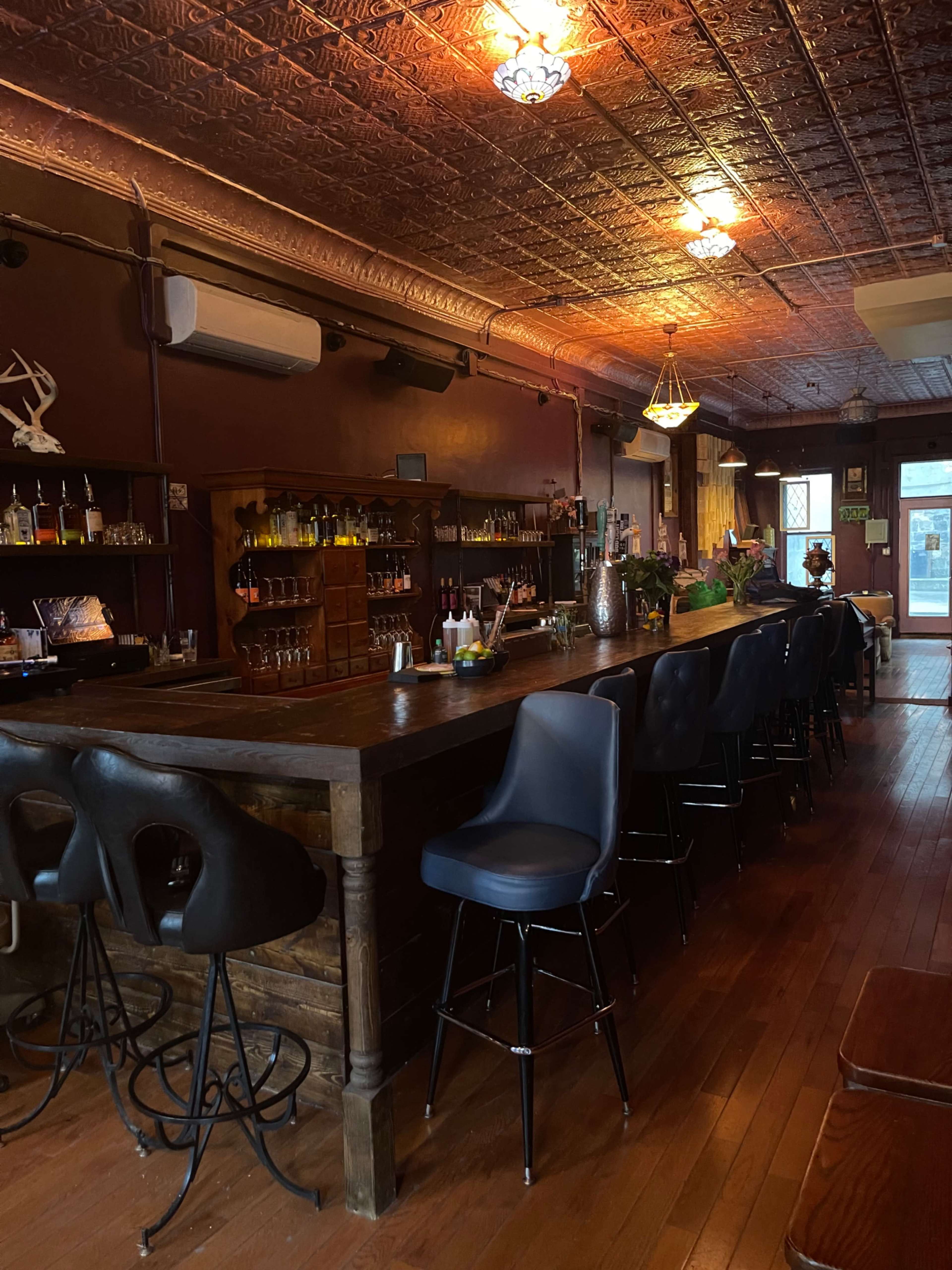 The image shows a wooden bar area in a dimly lit establishment with high stools and shelves stocked with various bottles.