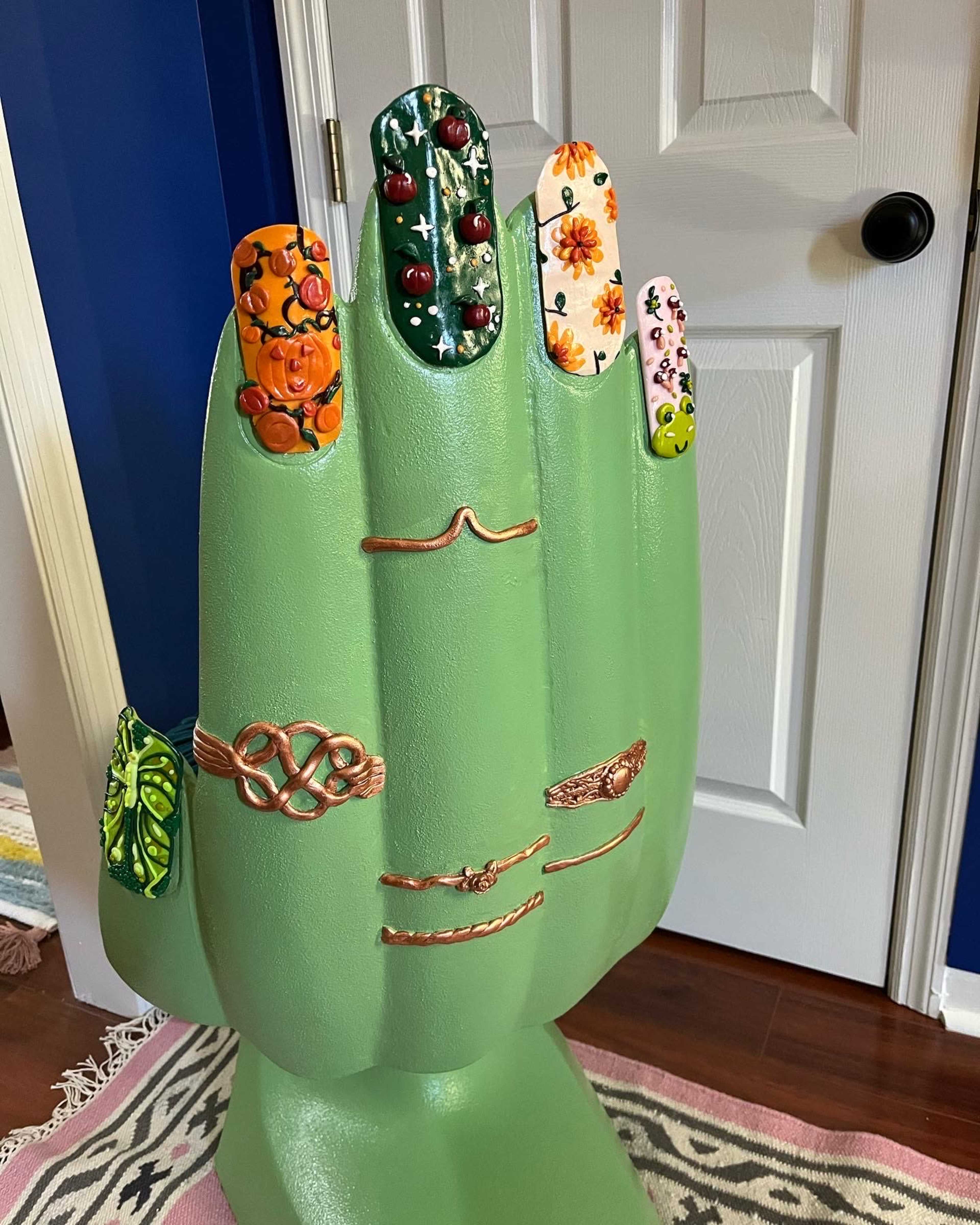 A large, green hand sculpture displays multiple painted fingernails decorated with various floral and artistic designs.