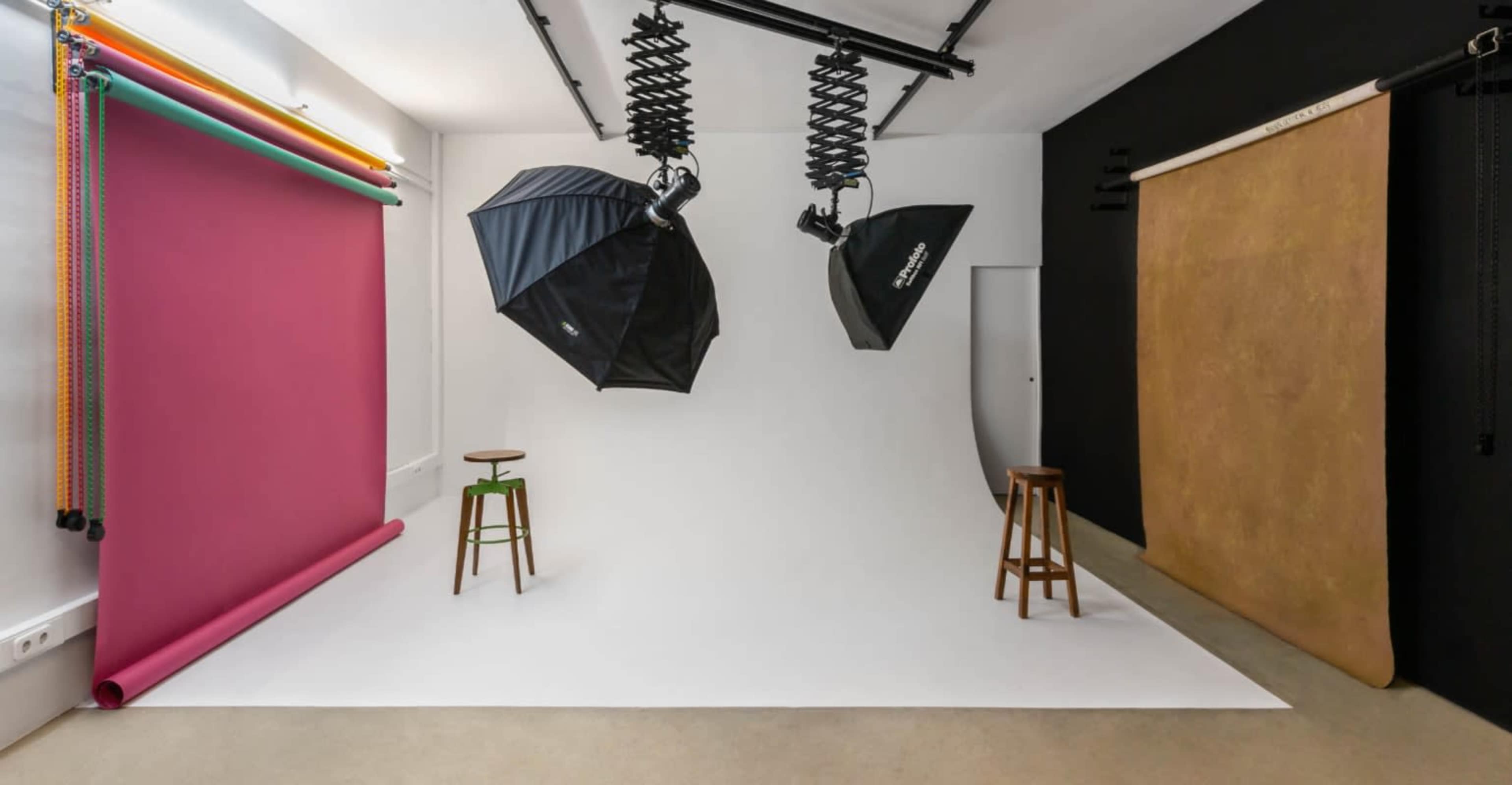 The image shows a photography studio with a white backdrop, two softbox lights, and colored paper rolls along a wall.