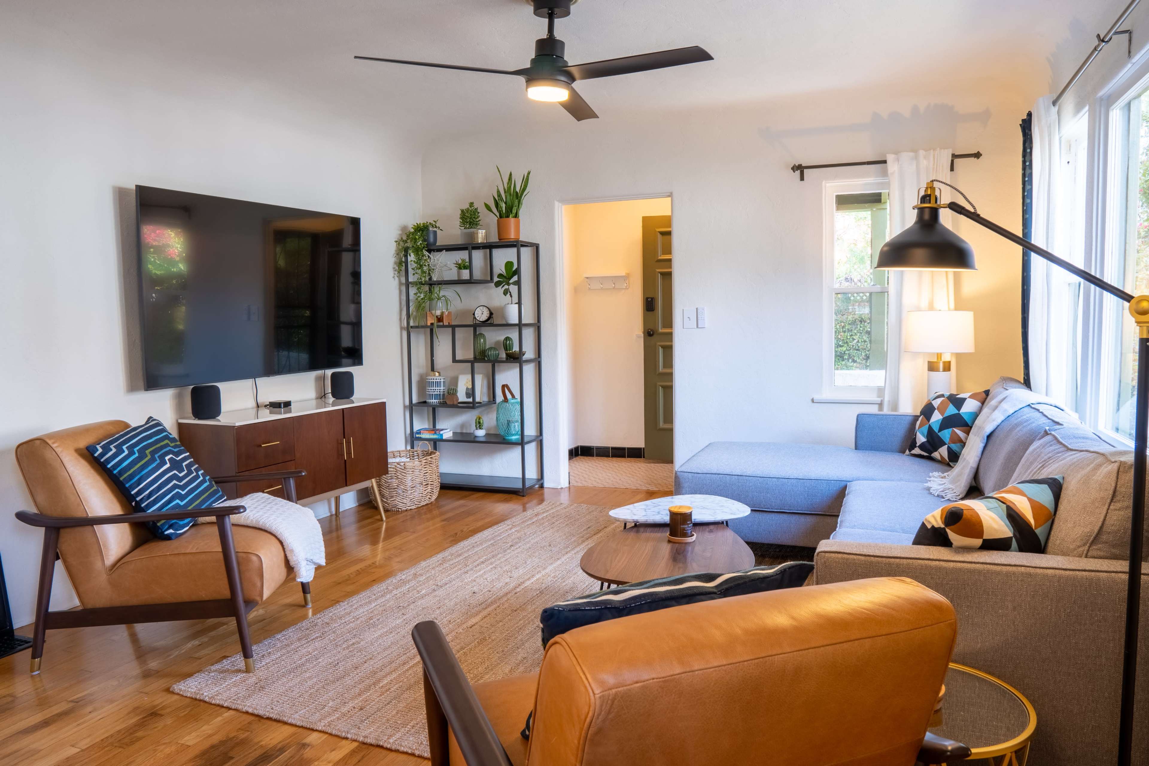The living room features a sectional sofa, two armchairs, a coffee table, a television mounted on the wall, and a shelf adorned with plants and decorative items.