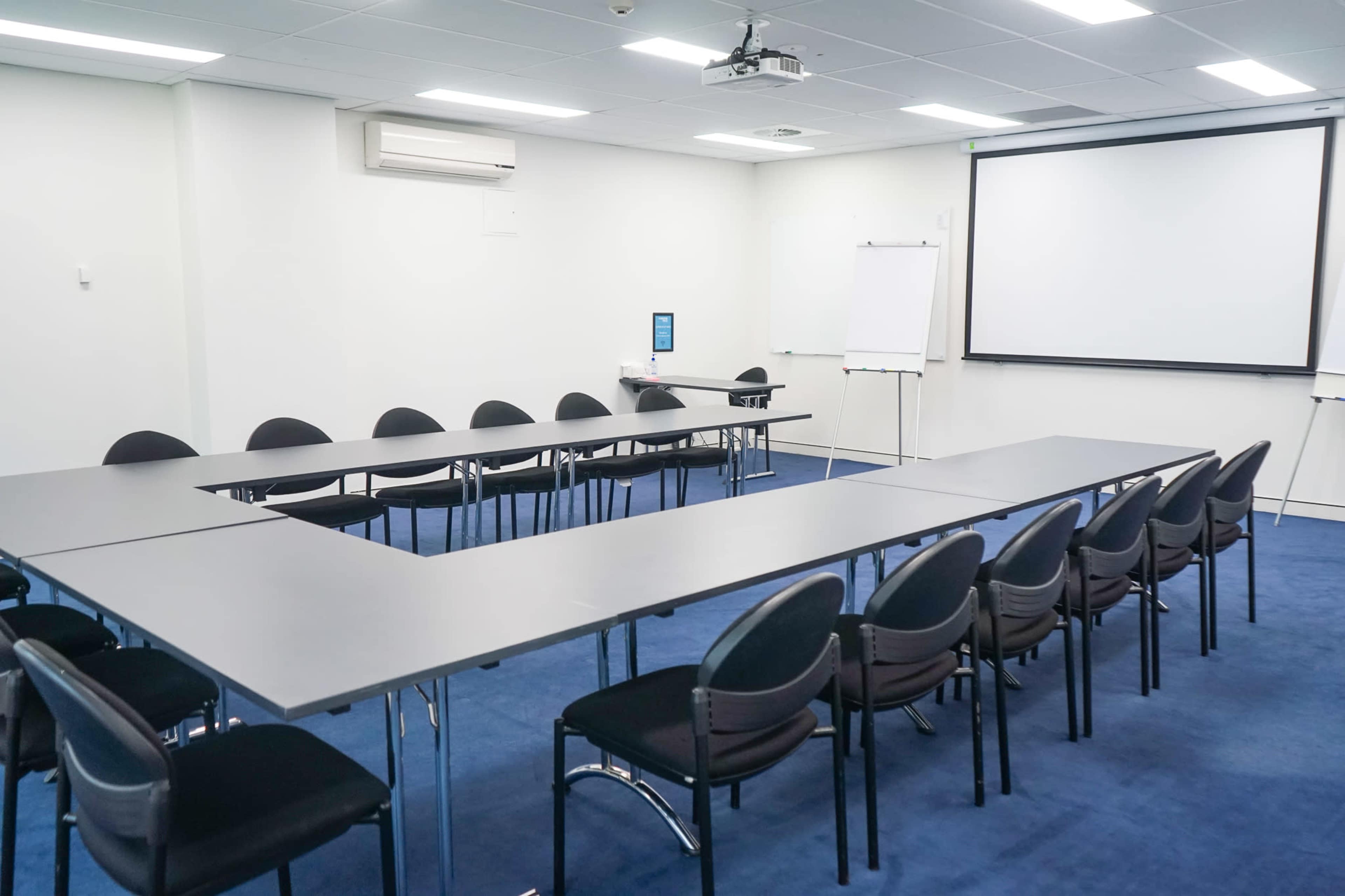 A conference room features a U-shaped arrangement of dark tables and black chairs, with a projector screen and whiteboard at one end.