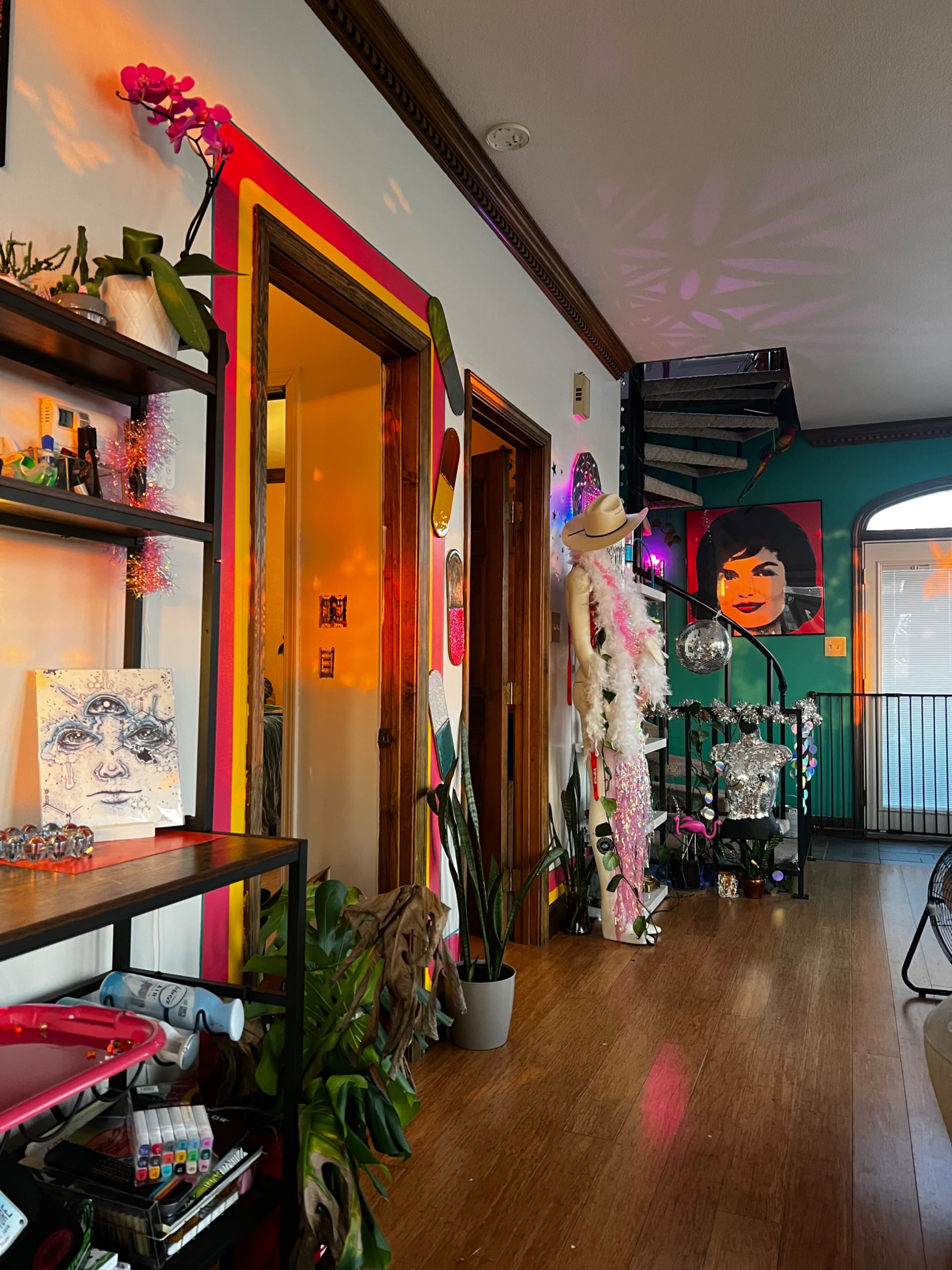 The image shows a colorful interior space featuring a staircase, a mannequin dressed in vibrant attire, and various decorative elements along the walls and shelves.