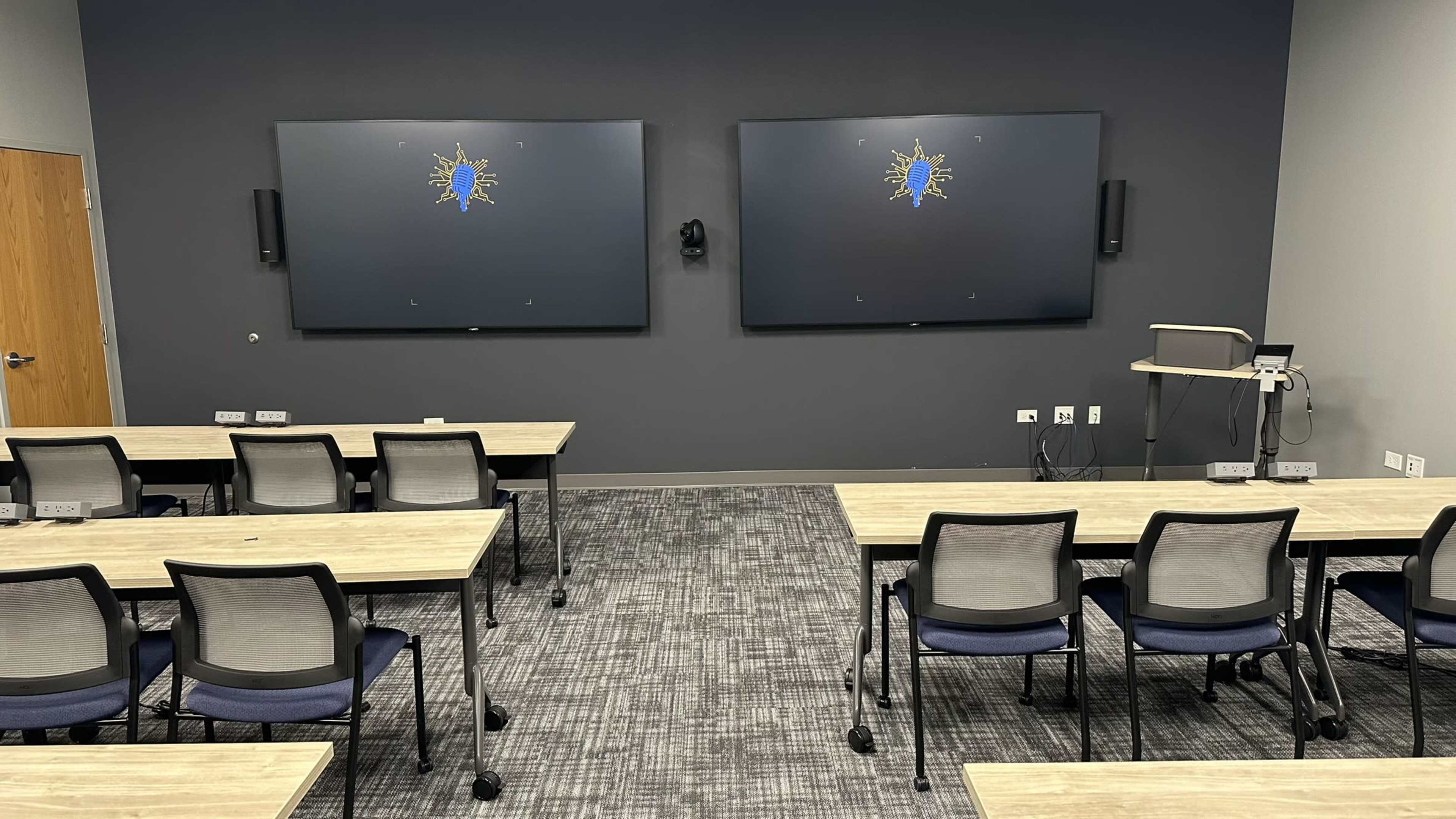 The image shows a modern classroom with two large screens on the front wall and several rows of tables and chairs arranged for instructional purposes.