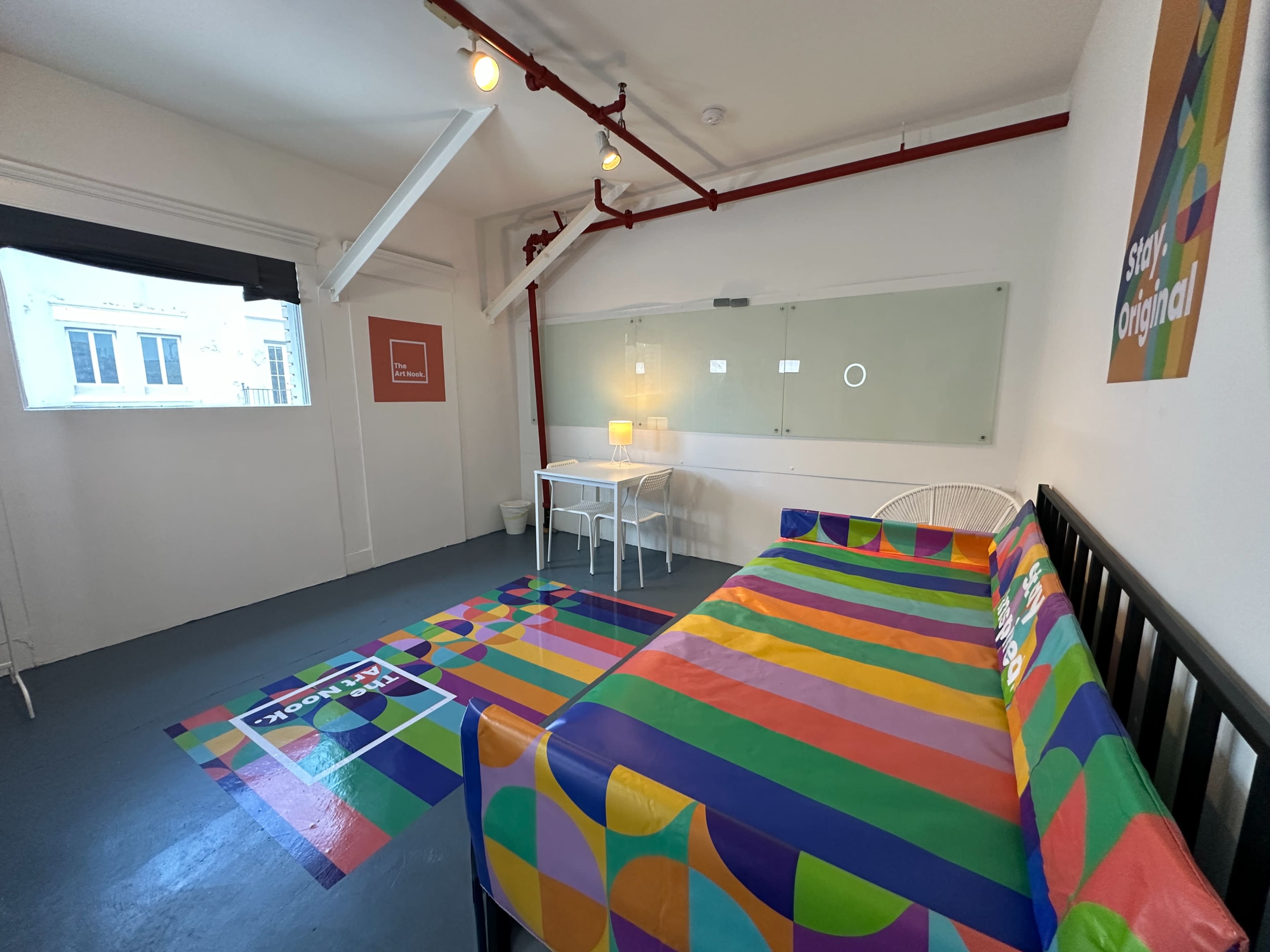 The image shows a brightly colored room with a striped bedspread, a small table and chair, and large windows letting in natural light.