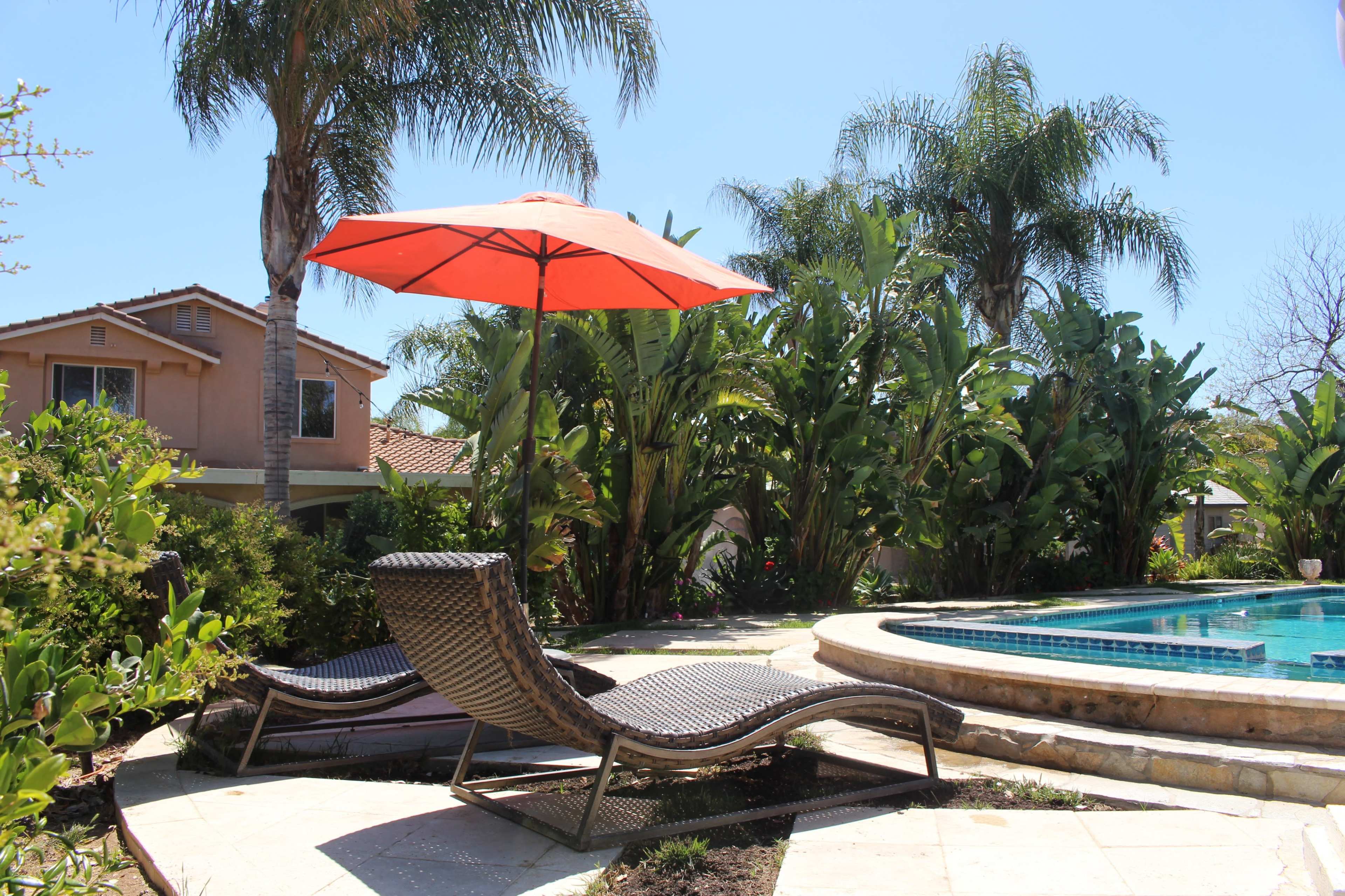 A poolside area features two lounge chairs beneath a large orange umbrella, surrounded by tropical plants and a residential house in the background.