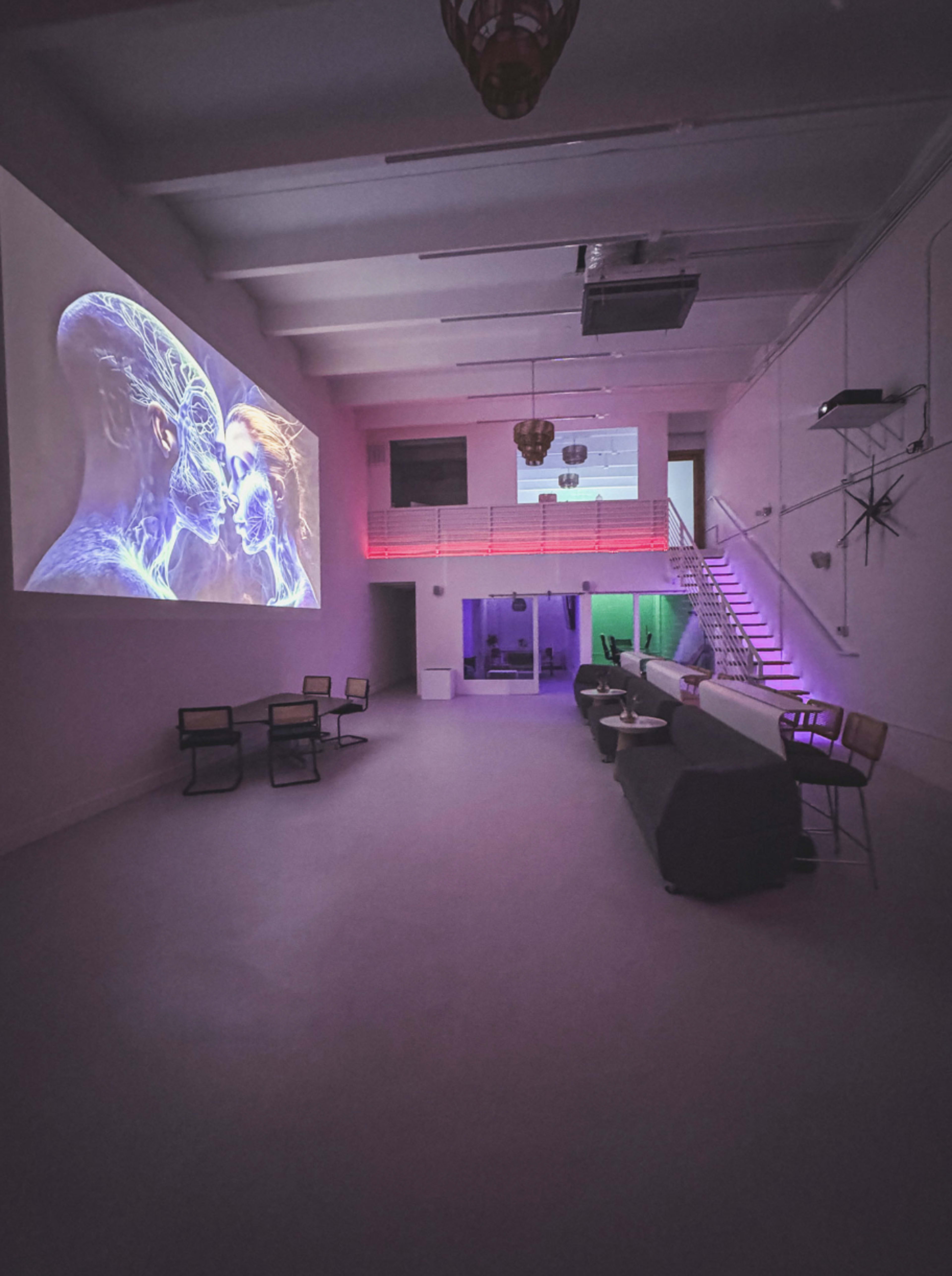The image shows a modern, spacious interior with a large projection on the wall, seating areas, and colorful lighting that enhances the minimalist design.