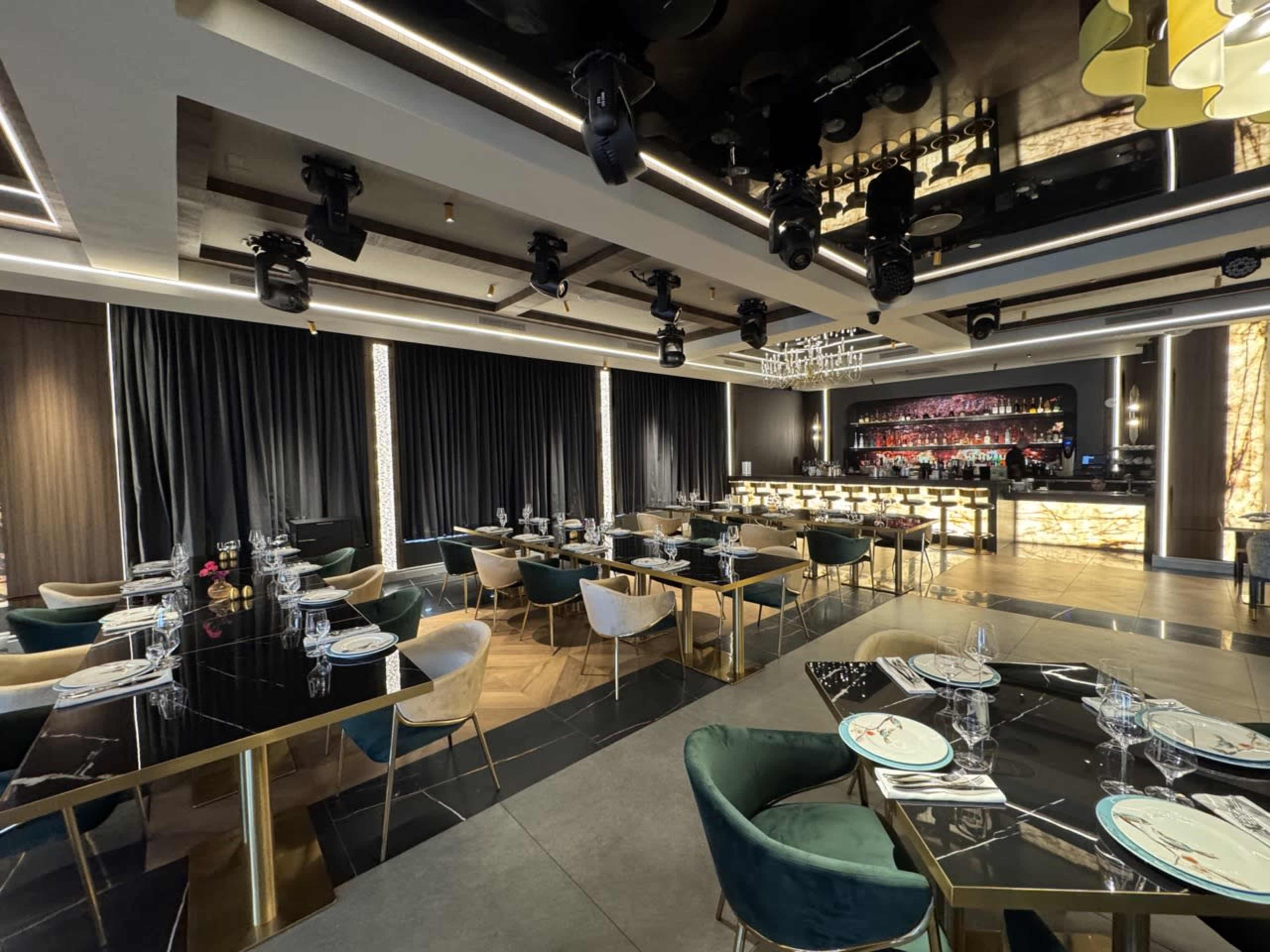 The image shows a modern restaurant interior with dark curtains, elegant dining tables, and a well-lit bar area in the background.