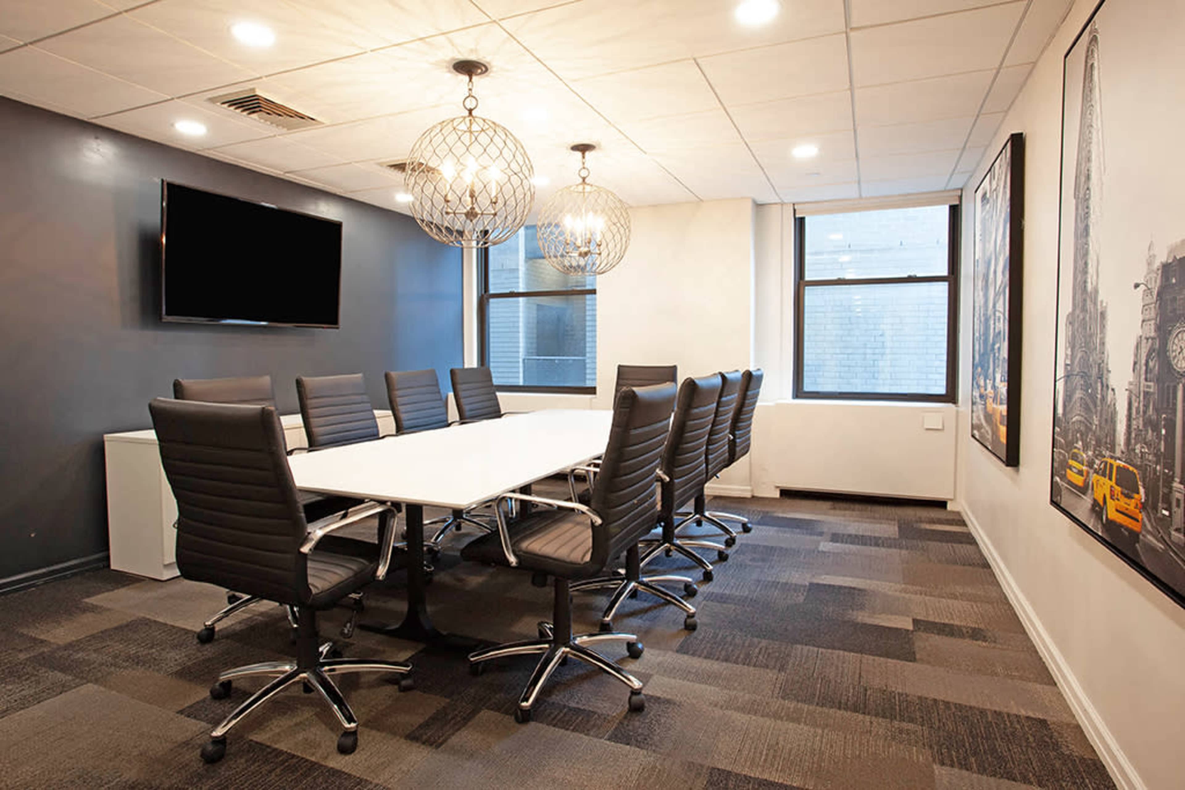 A modern conference room features a long white table surrounded by black swivel chairs, with pendant lighting and a wall-mounted TV.