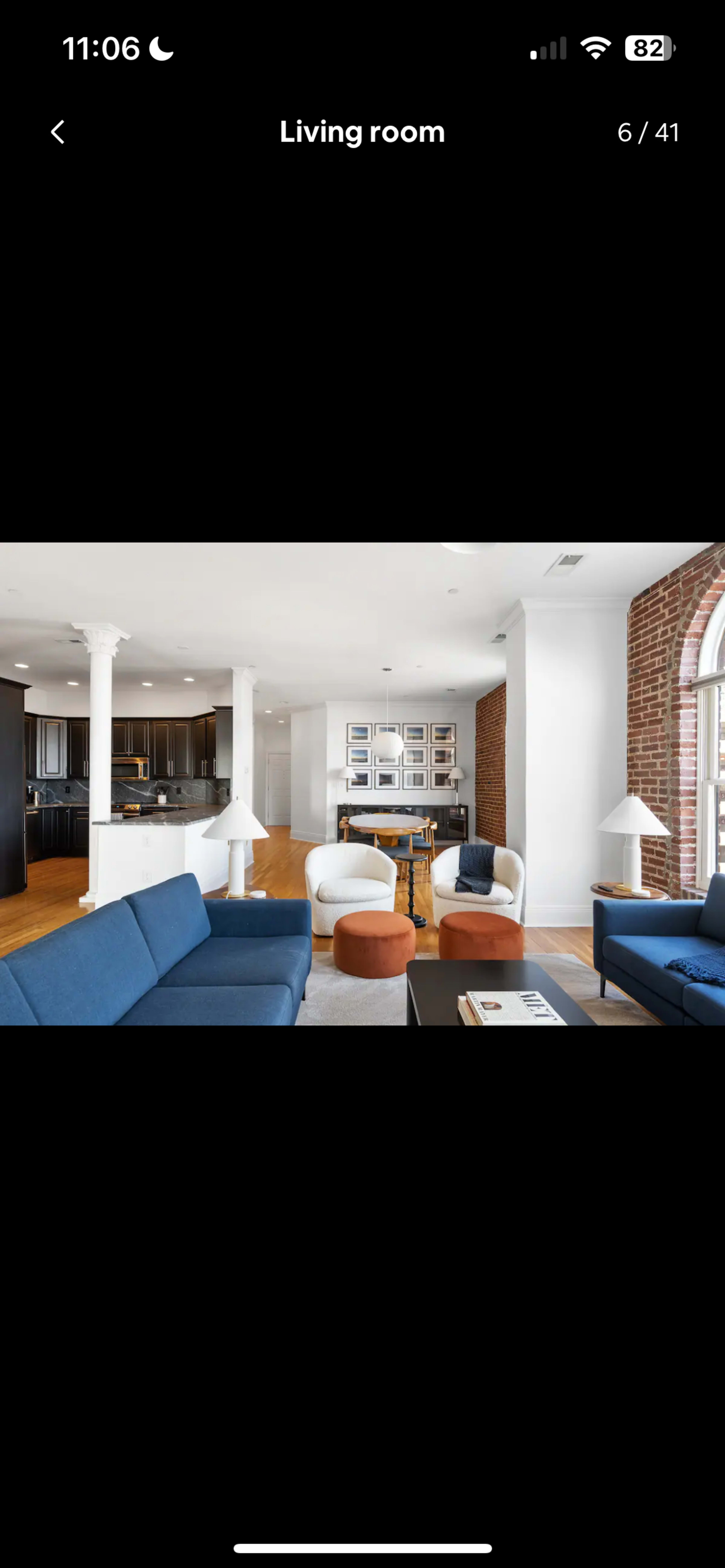The image shows a modern living room with a blue sectional sofa, round ottomans, and a view of a kitchen in the background.