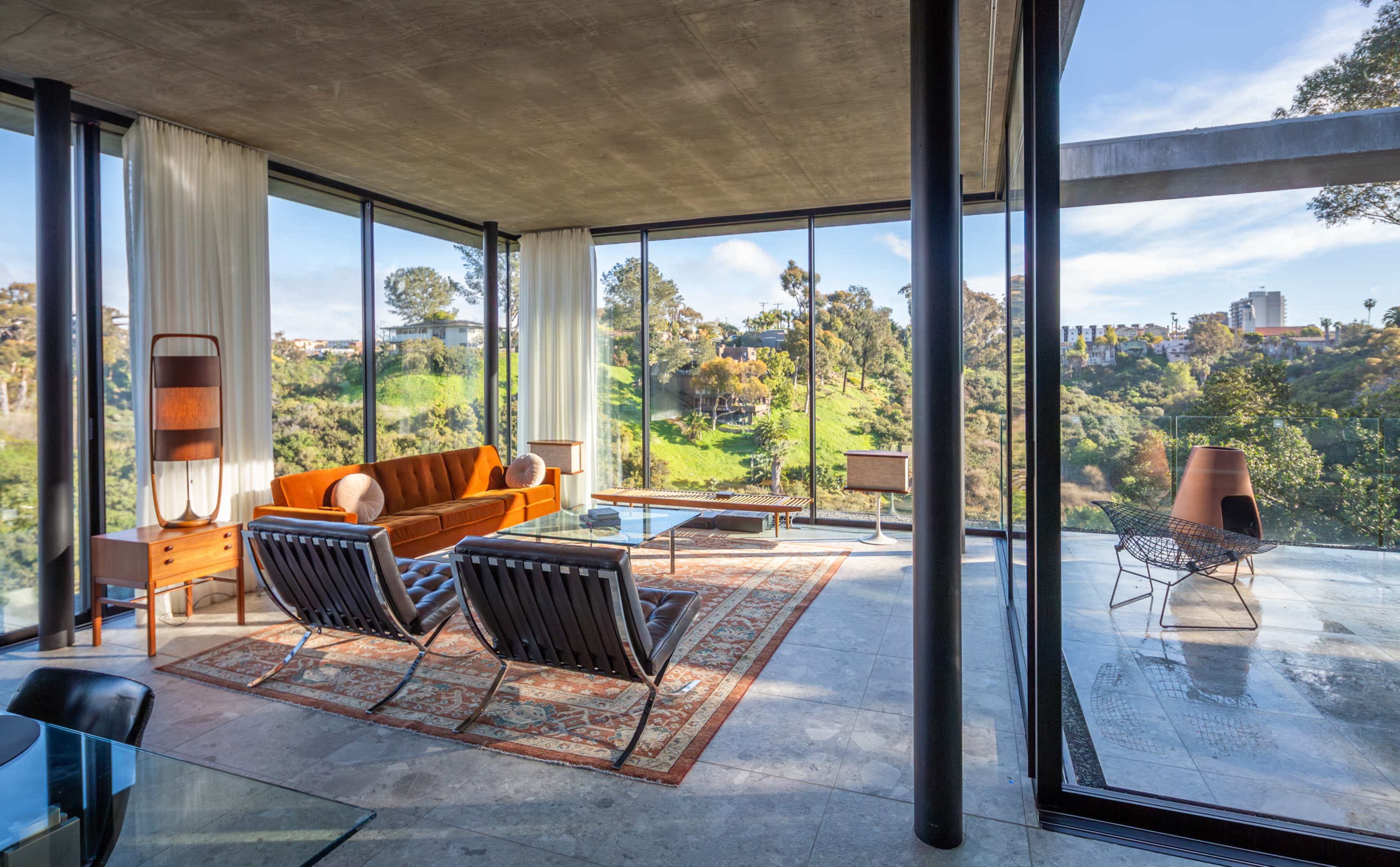 The image shows a modern living space with large glass windows, featuring an orange sofa, two black chairs, and a view of green hills outside.