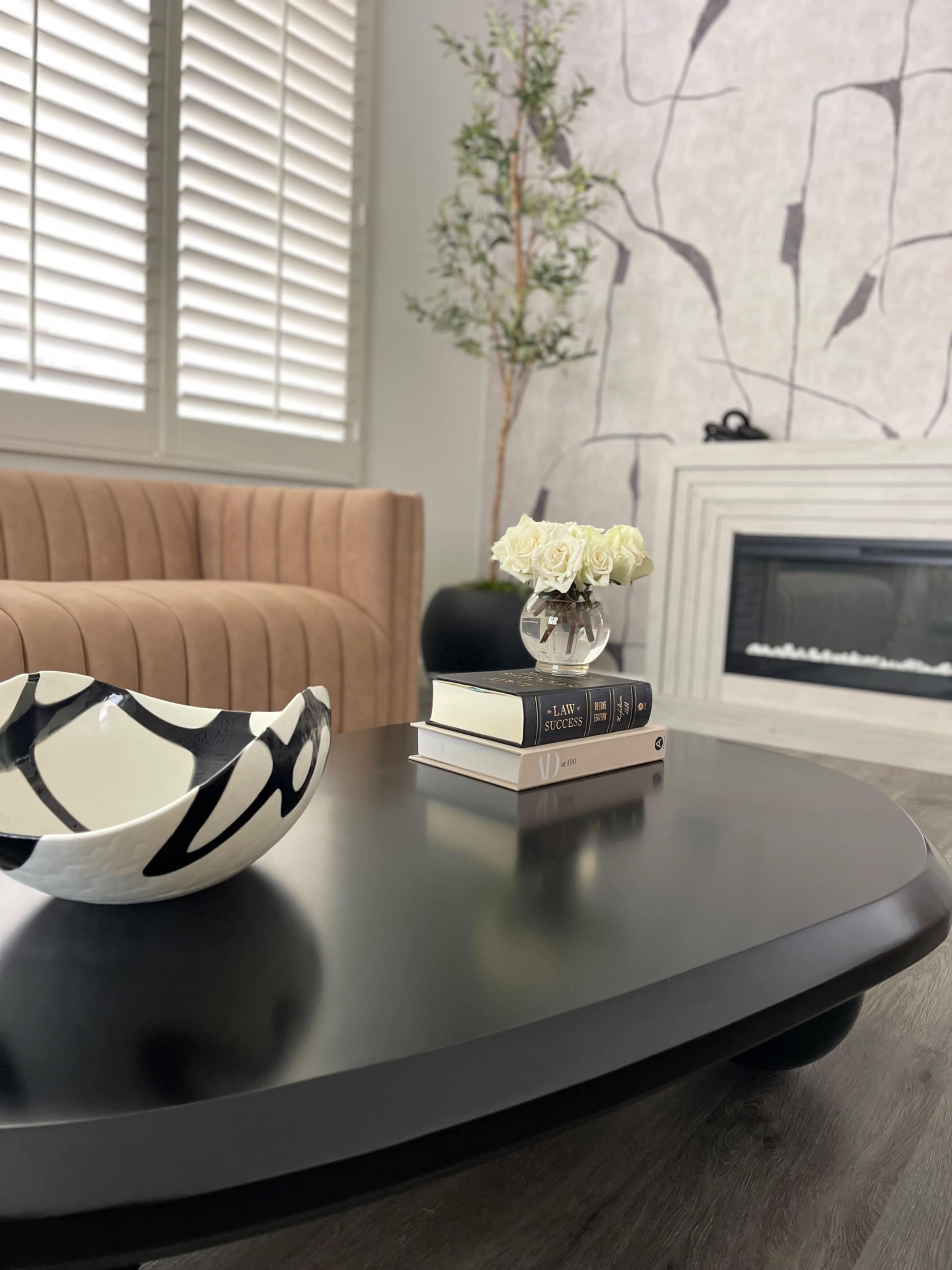 A modern living room features a beige sofa, a black coffee table with decorative books and a bowl, a small vase of white roses, and a fireplace against a textured wall.