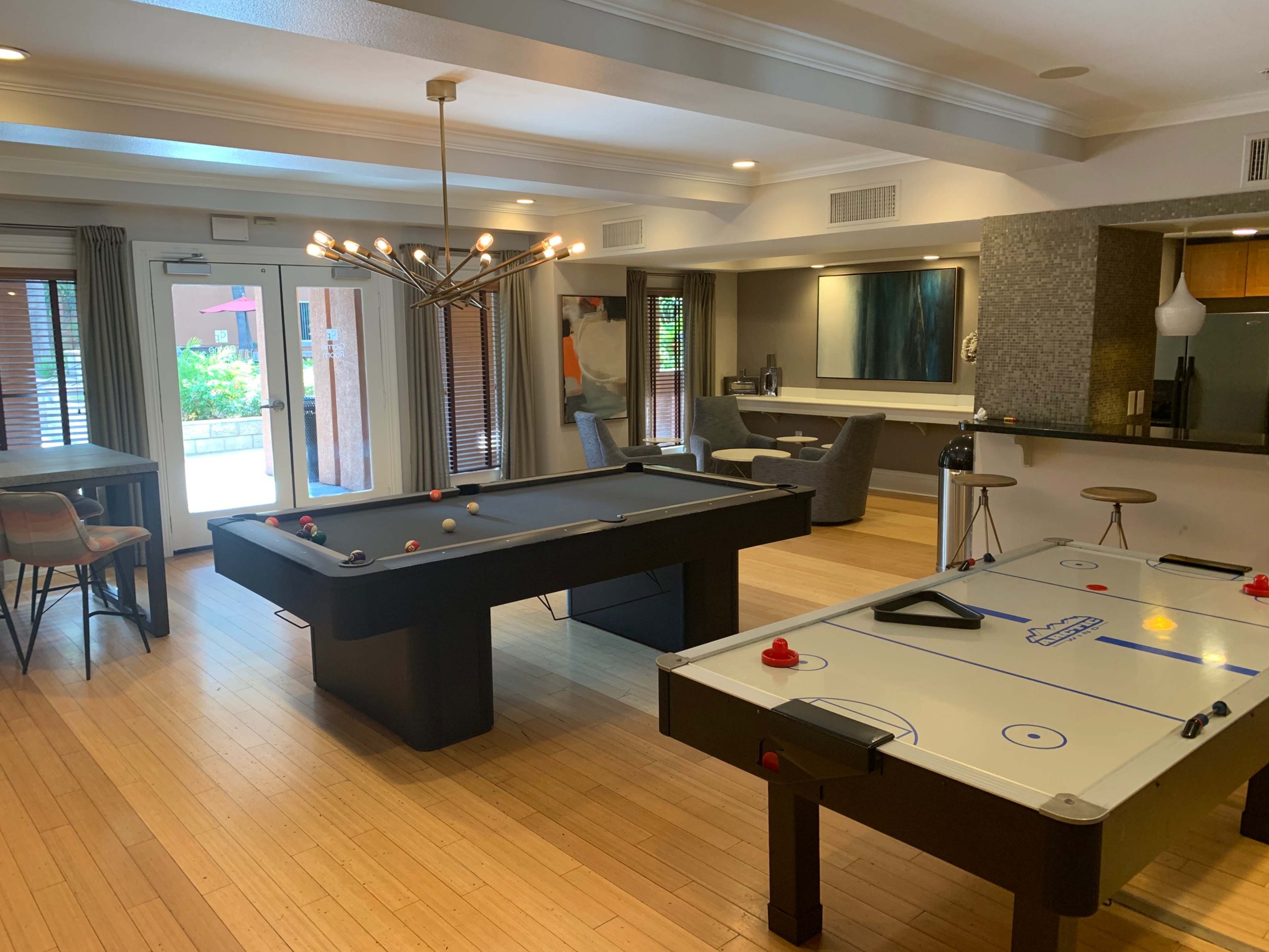 A spacious game room featuring a pool table and an air hockey table, with a dining area and kitchenette in the background.