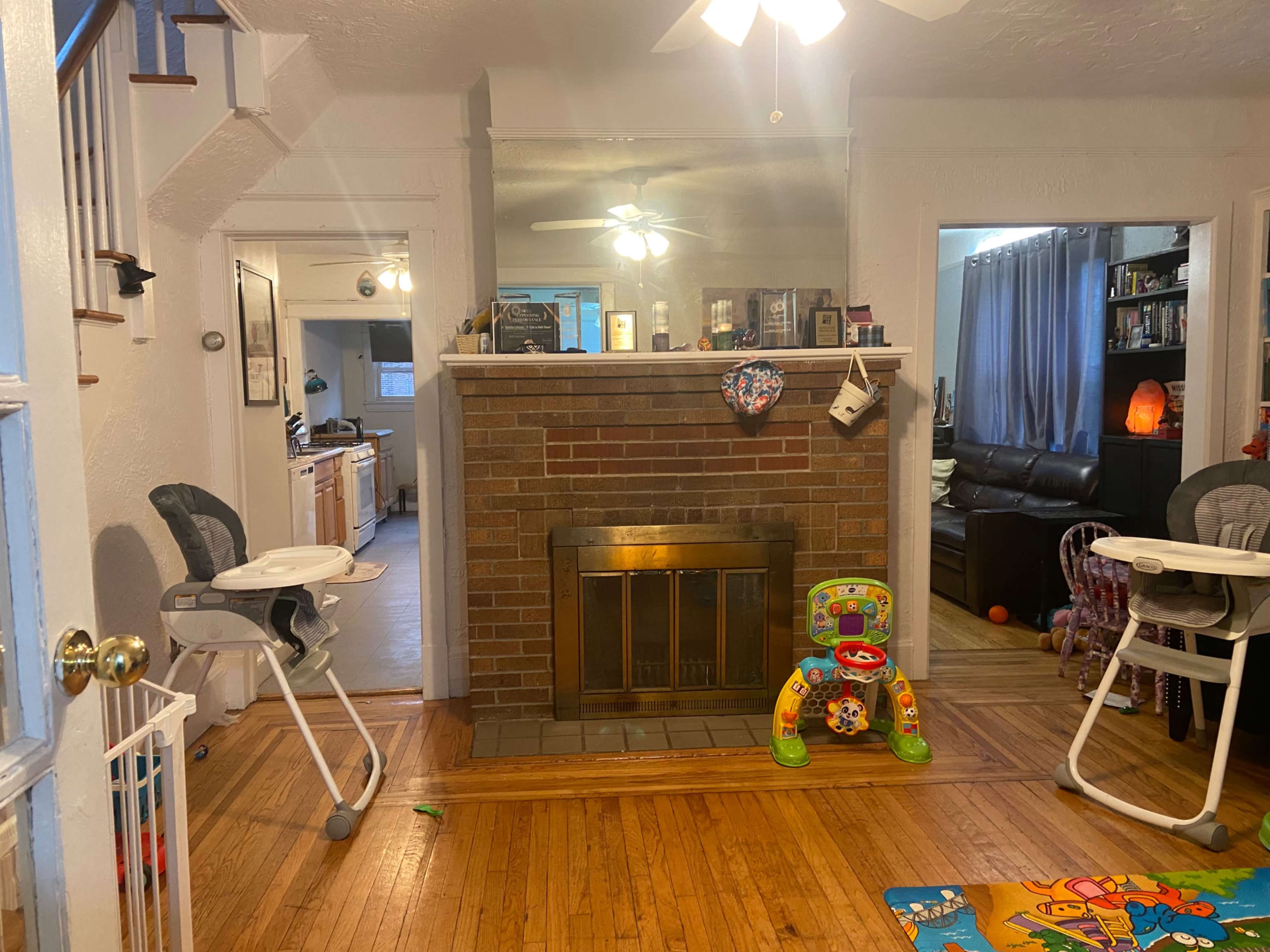 The image shows a living room with a brick fireplace, two high chairs, a colorful toy, and visible kitchen access in the background.