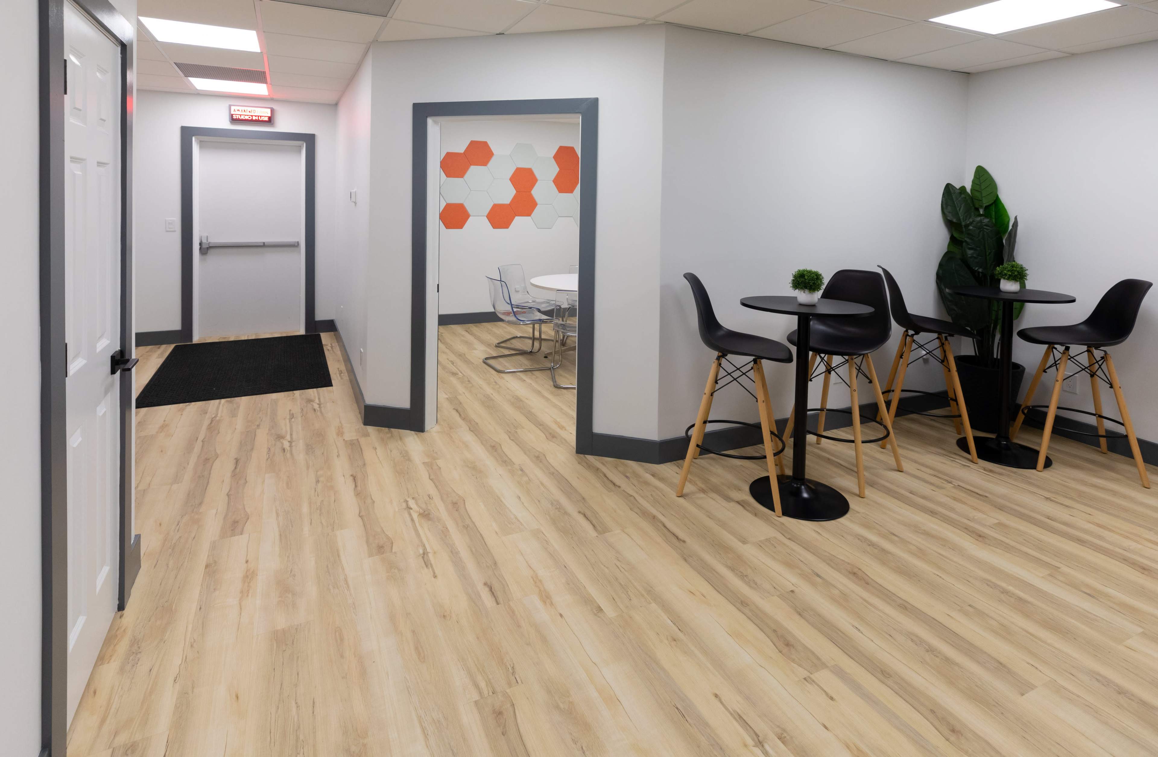 A modern office hallway with light wood flooring, leading to a break area with tall black chairs and a small round table beside a decorative plant.
