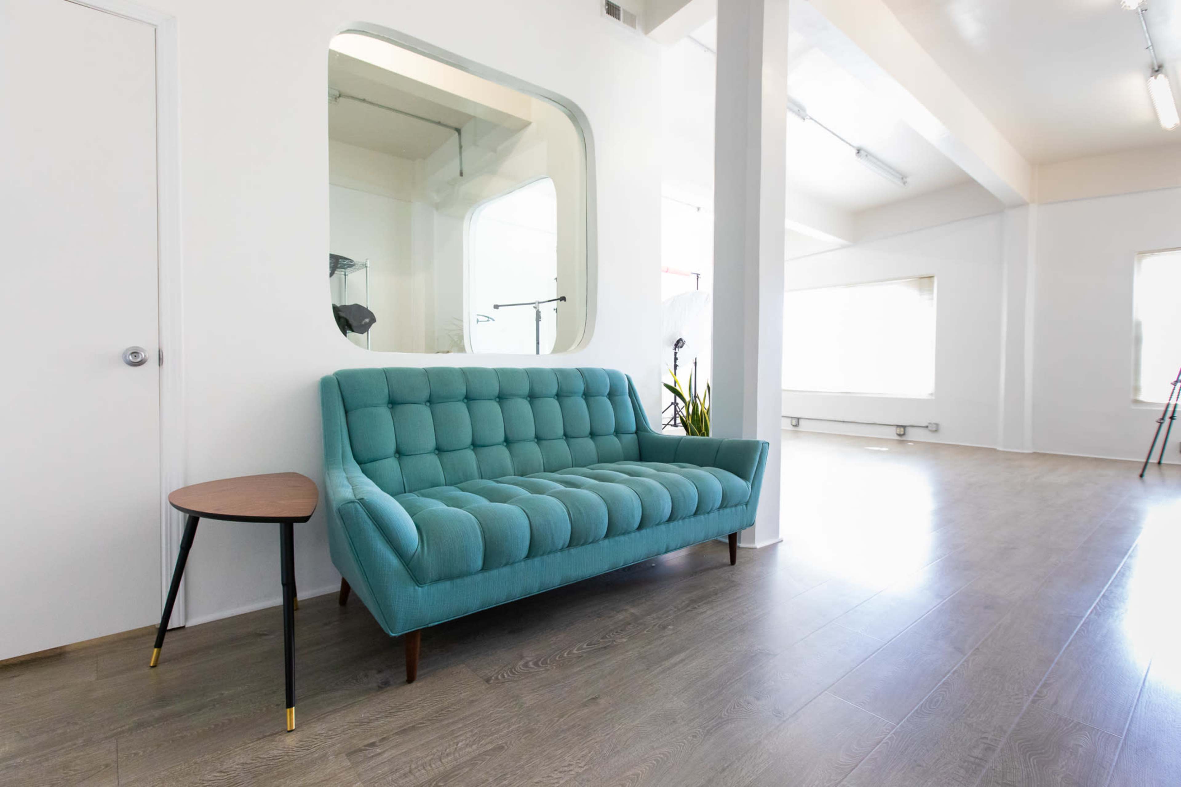 A light-filled room with a teal upholstered sofa and a small round side table next to it, featuring large windows and a minimalist design.