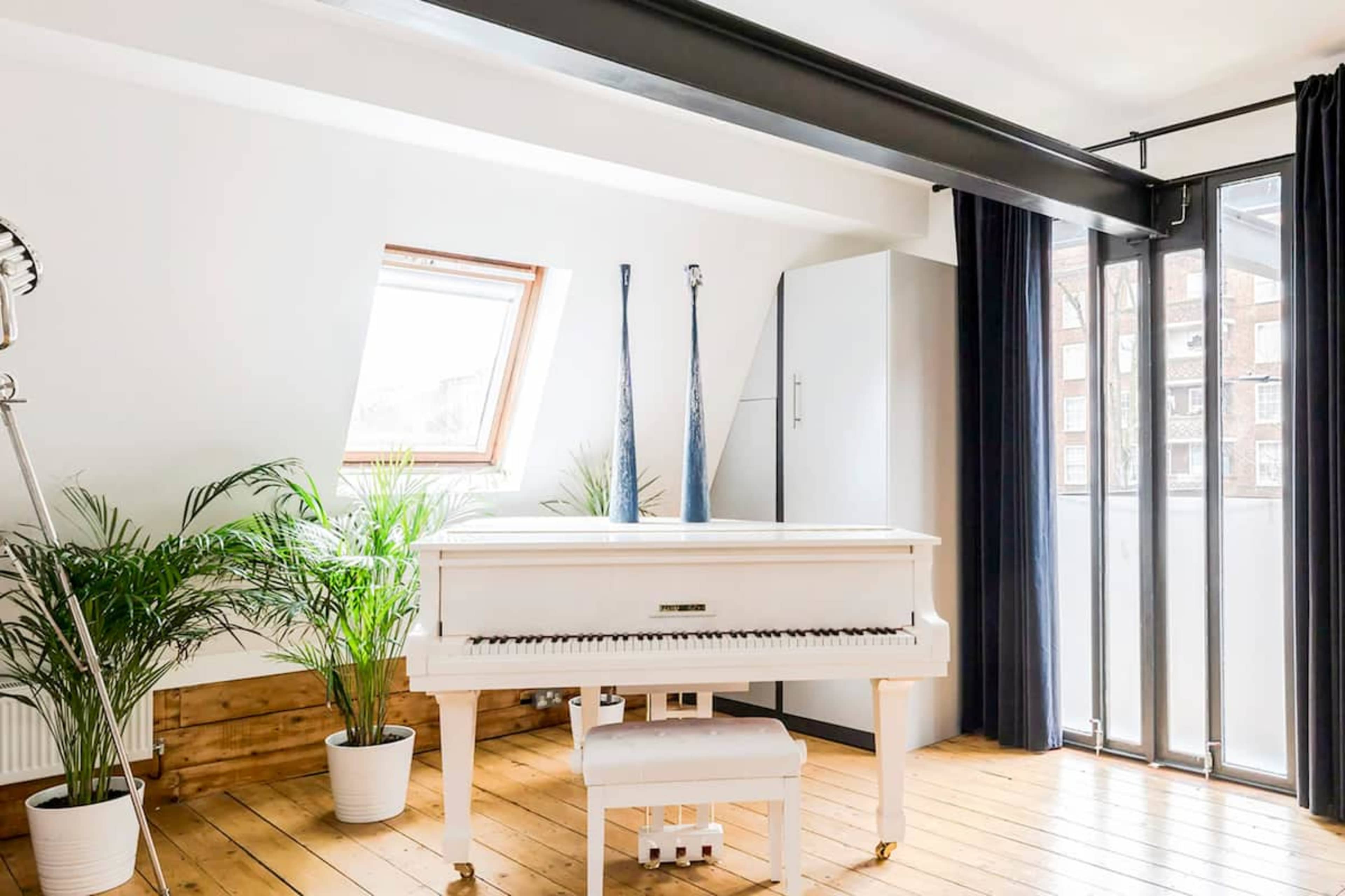 A white piano is positioned in a bright, airy room with wooden flooring, potted plants, and large windows.