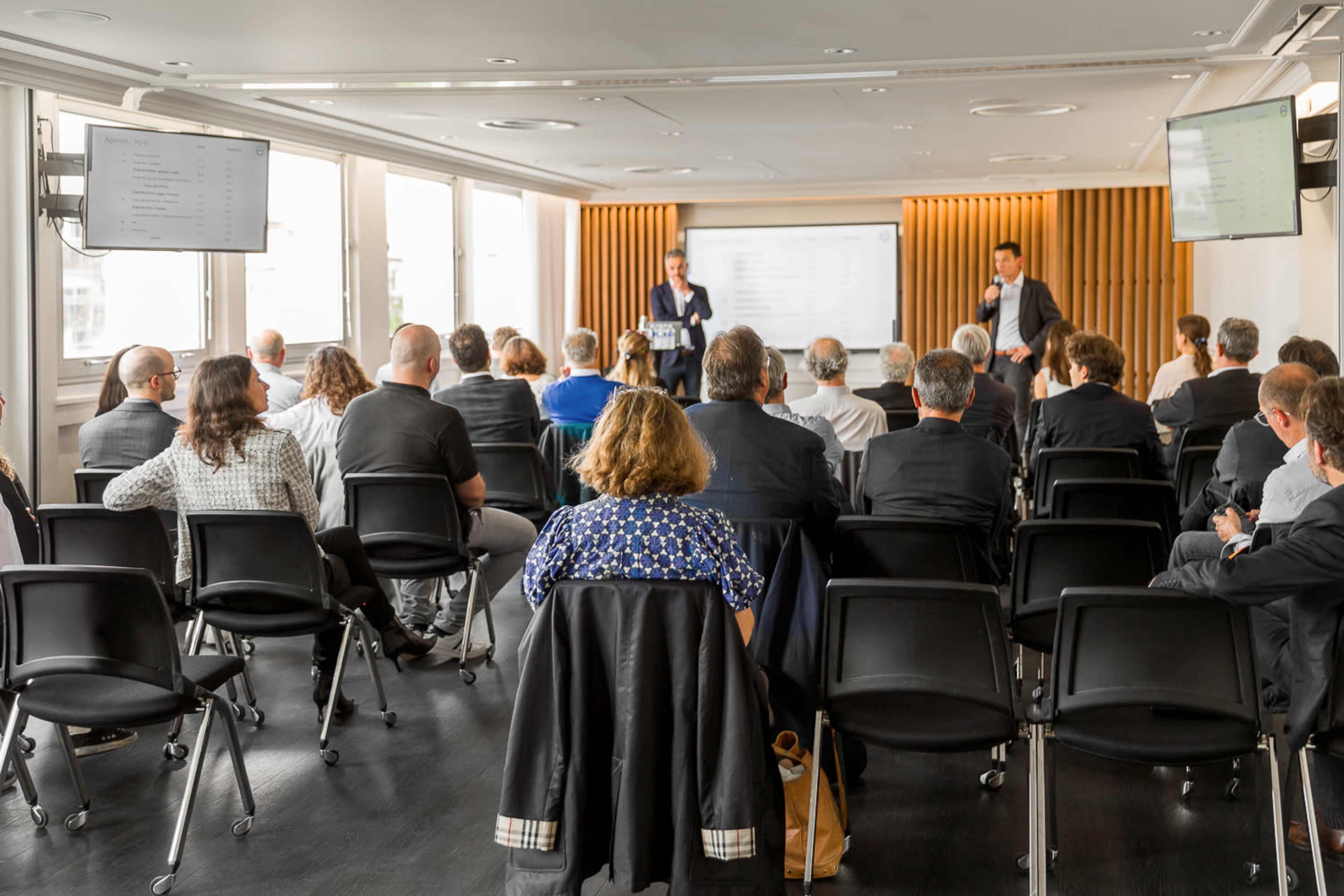A group of people sits in a conference room, facing two speakers presenting in front of a large screen.