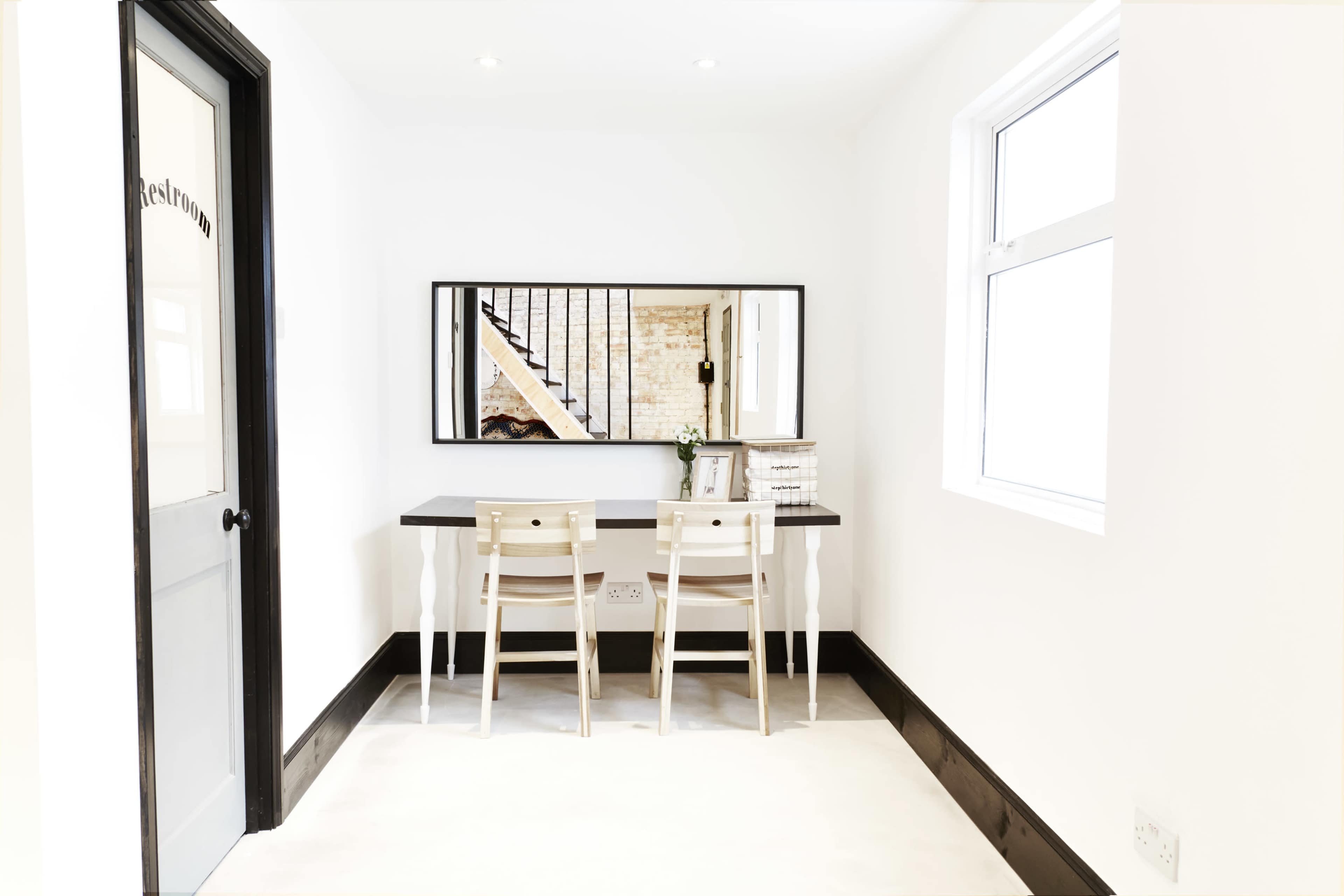 The image shows a minimalist room with a black and white color scheme, featuring a table with two chairs and a large mirror on the wall opposite a door labeled "Restroom."