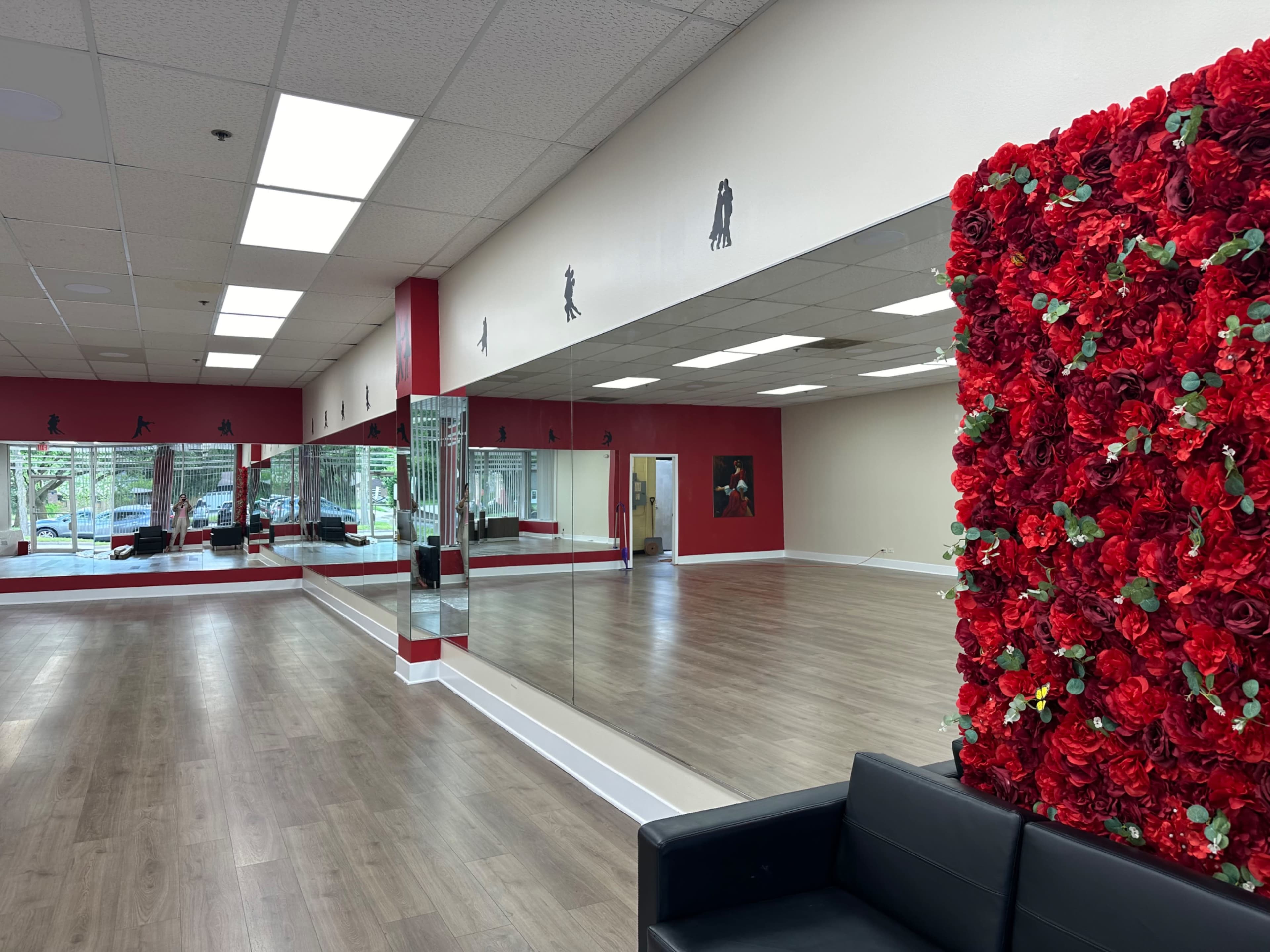 A dance studio with a mirrored wall, wooden flooring, and a vibrant floral accent wall near a black seating area.