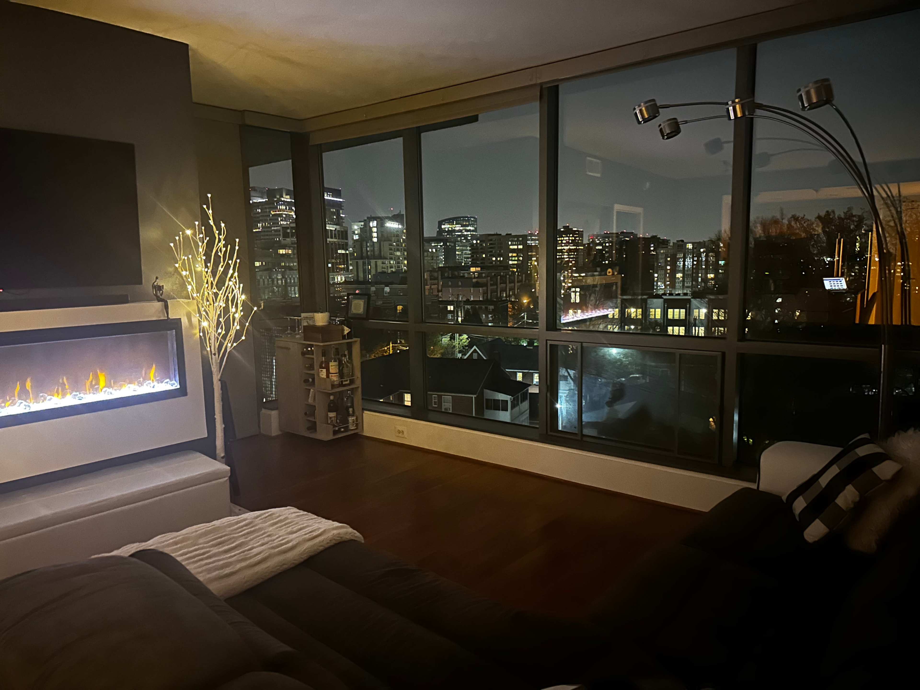 The scene shows a modern living room with a large window displaying a city skyline at night, featuring a fire-like electric heater and a decorative floor lamp.
