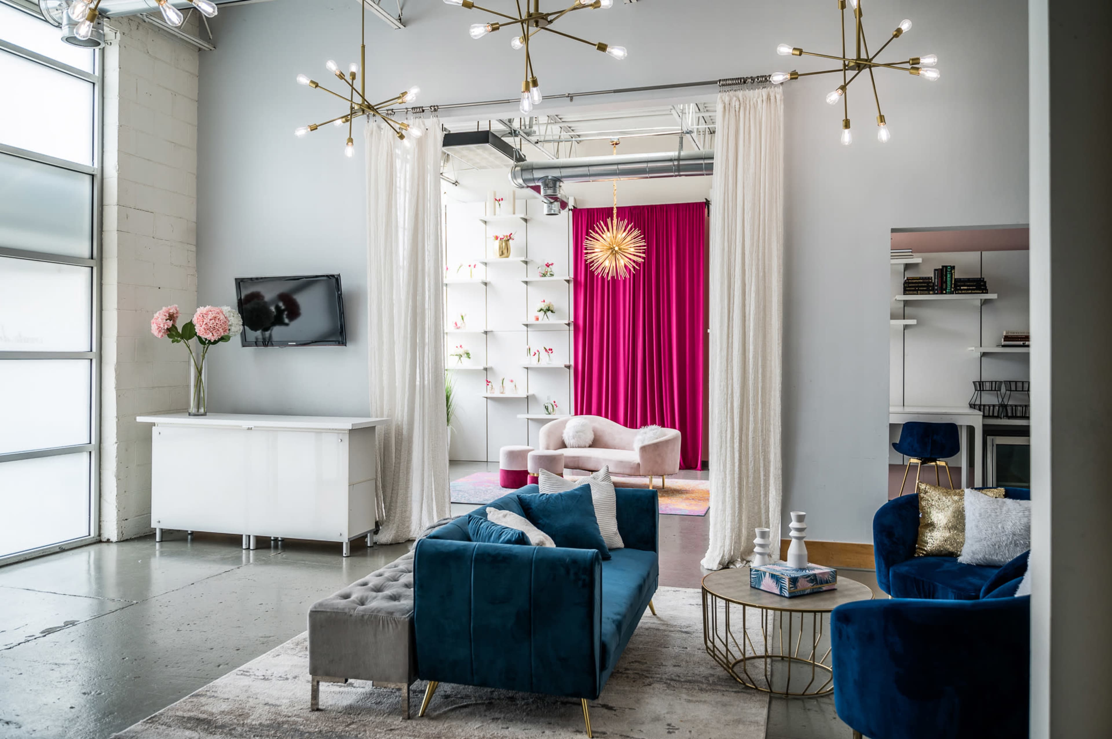 A modern interior space features a living area with a blue velvet sofa, a coffee table, and a white cabinet, with a backdrop of pink curtains and decorative shelves.