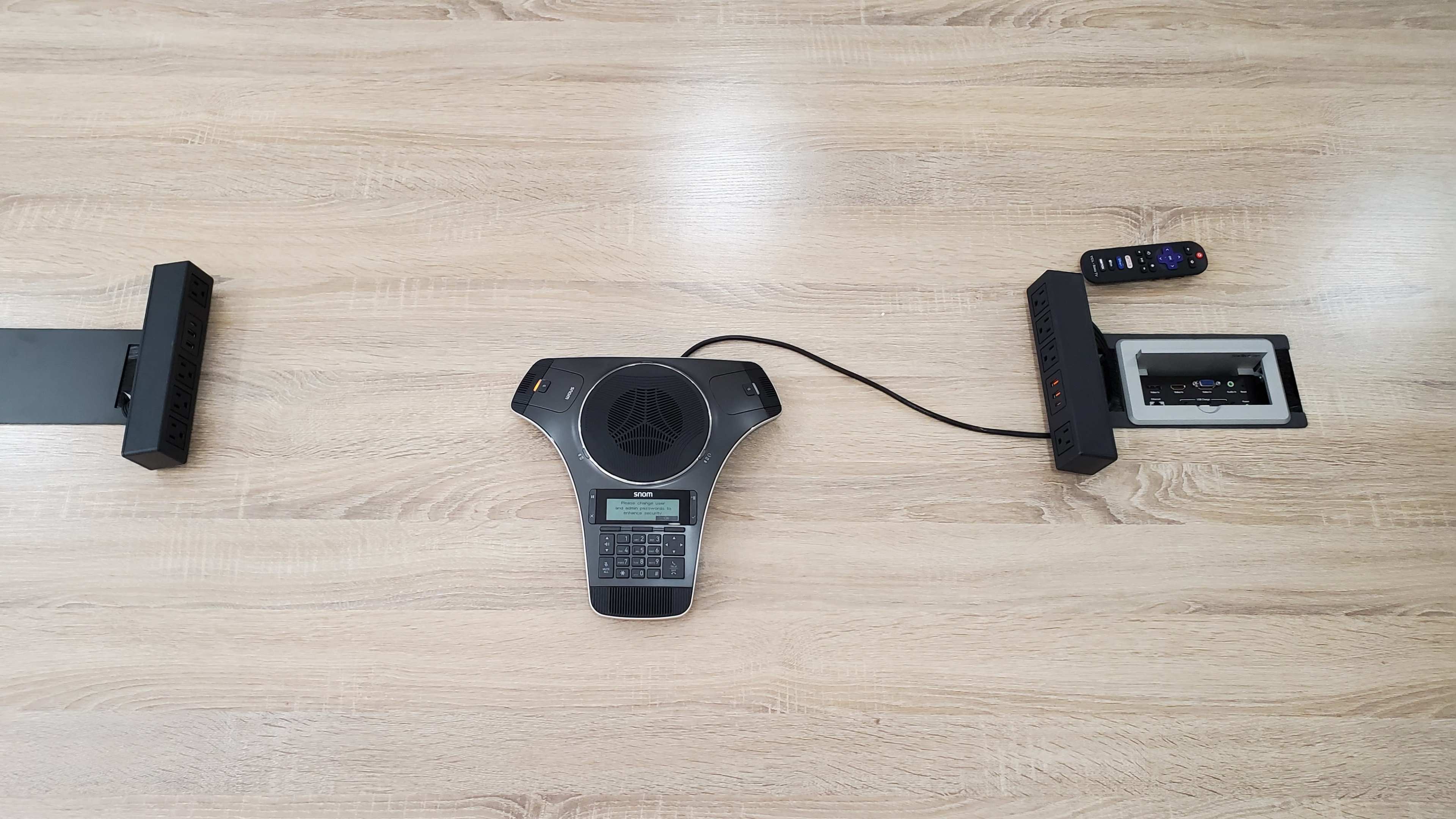 The image shows a conference table with a speakerphone and remote control placed centrally, flanked by two black device mounts.