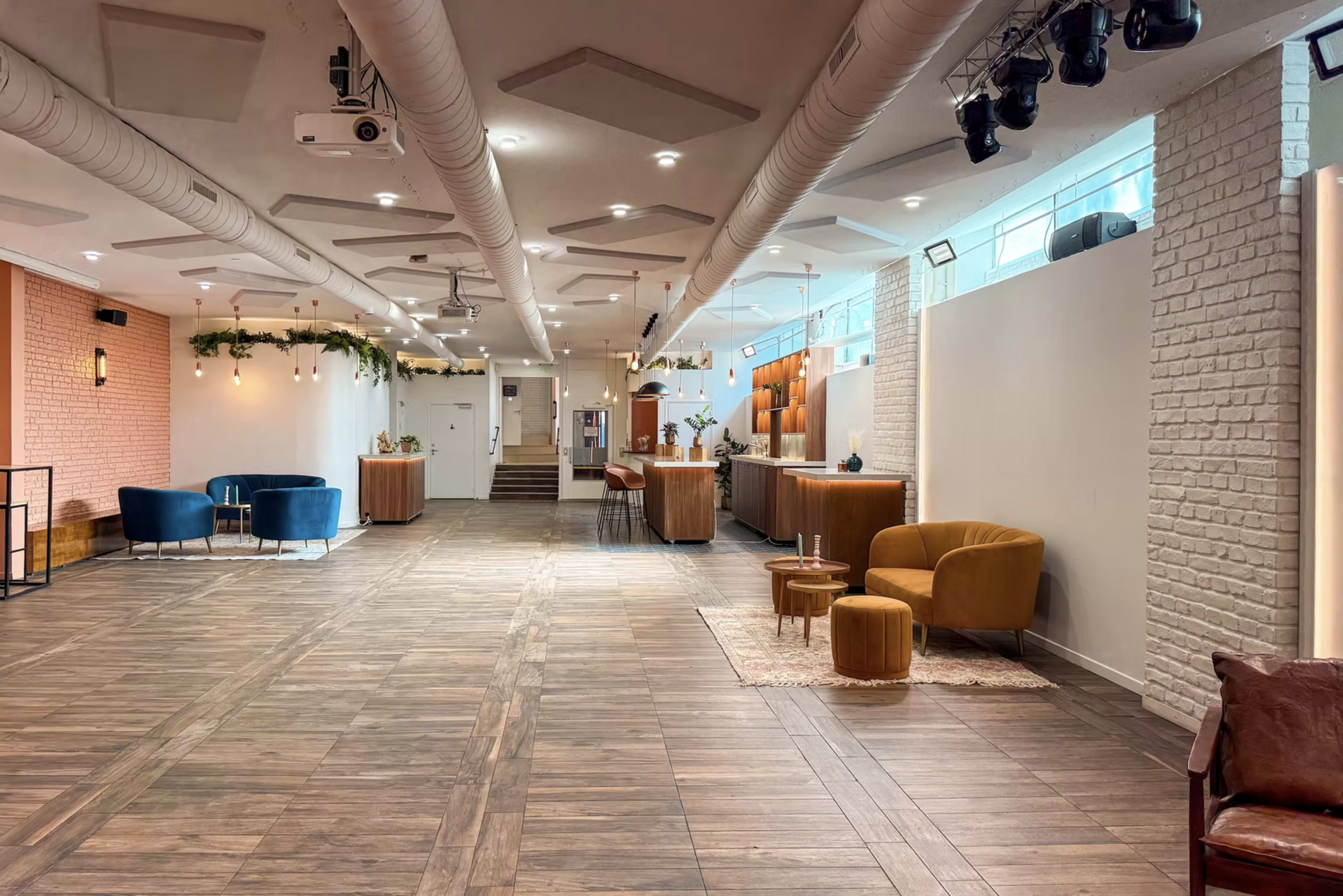 The image shows a spacious interior with a wooden floor, featuring seating areas with blue and orange chairs, a bar counter, and decorative plants along the walls.