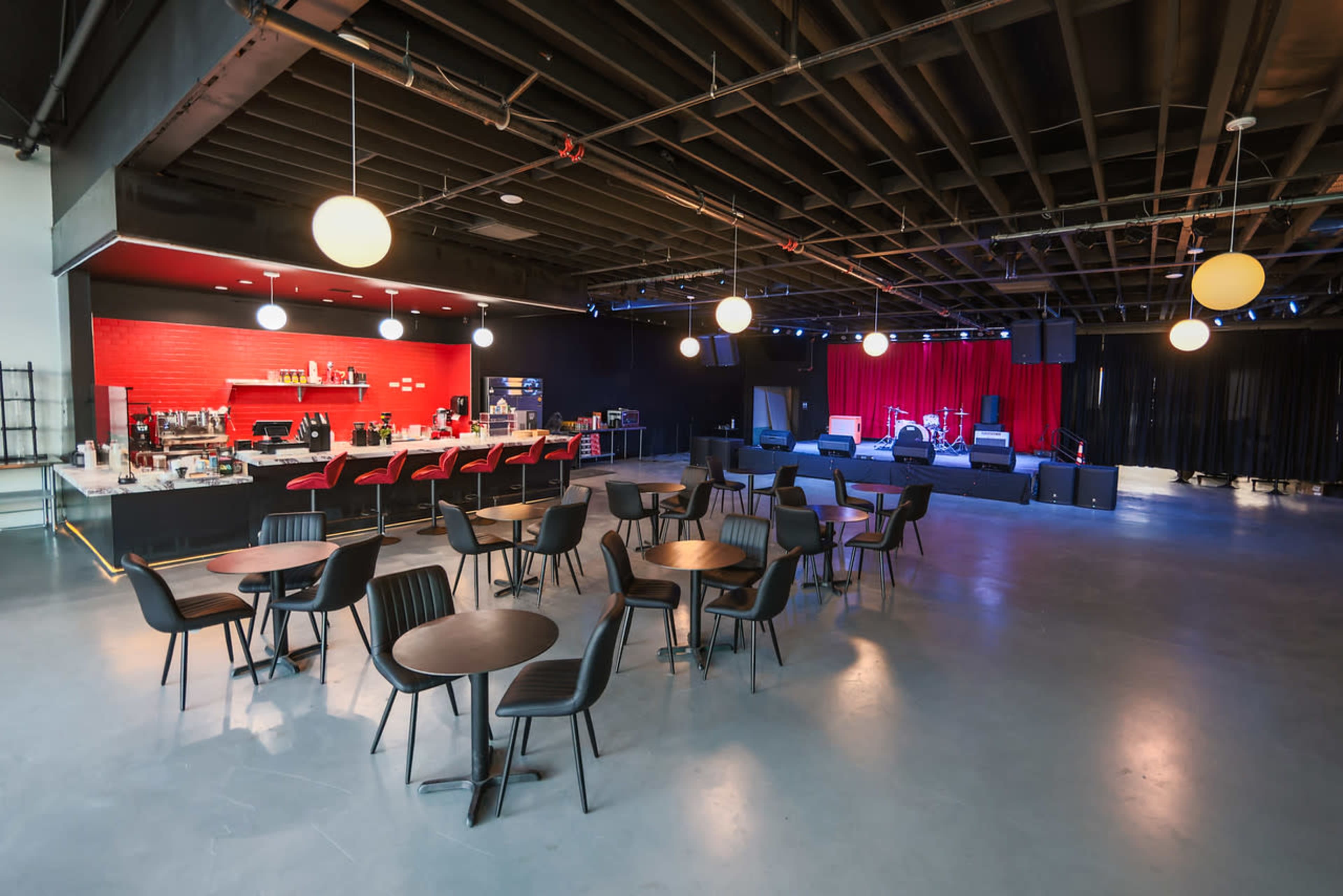 The image shows a spacious indoor venue with black tables and chairs set up, a red bar area in the background, and a stage with red curtains and lighting.