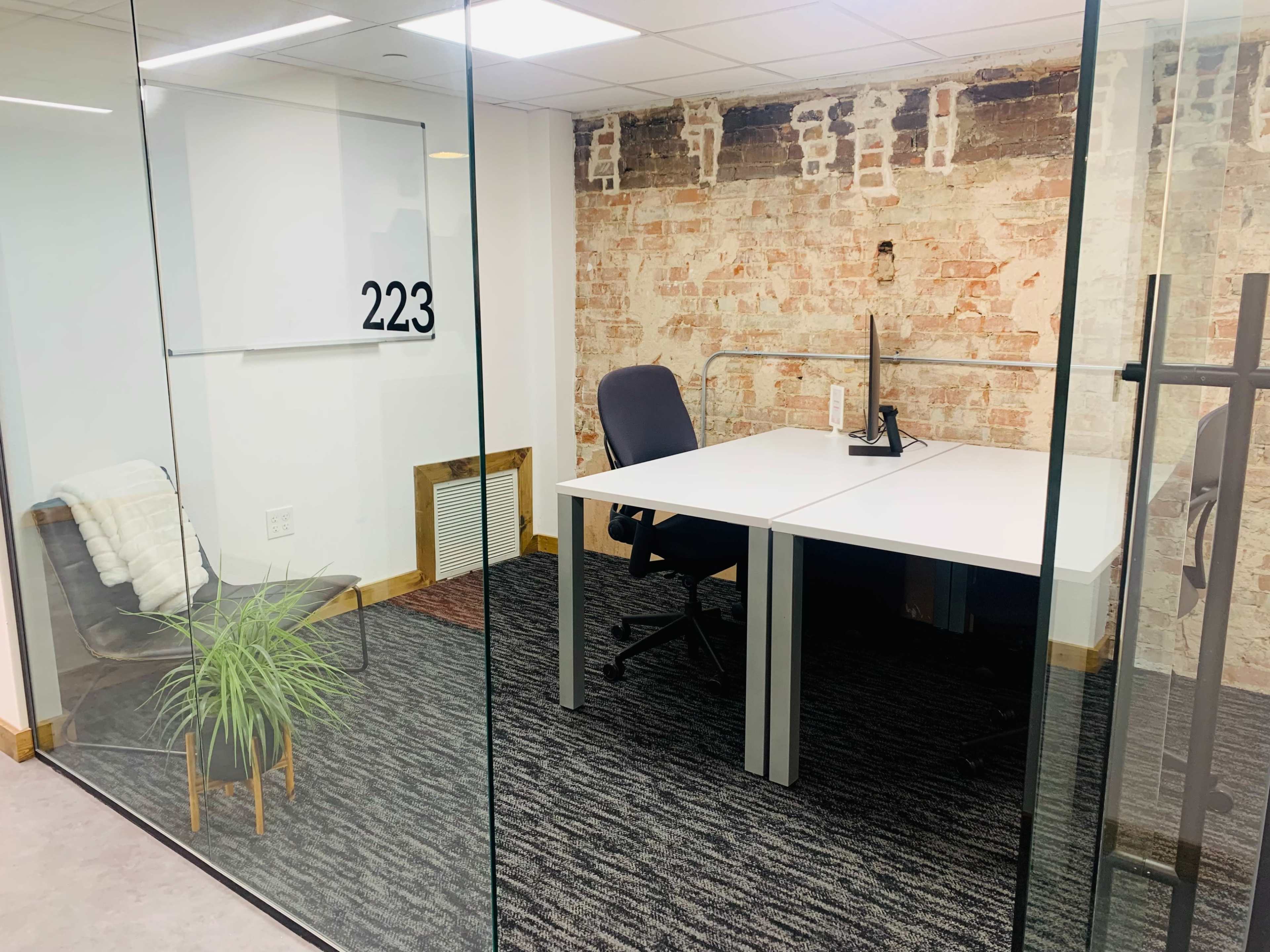 An office space numbered 223, featuring a white desk, a chair, a plant, and exposed brick walls.
