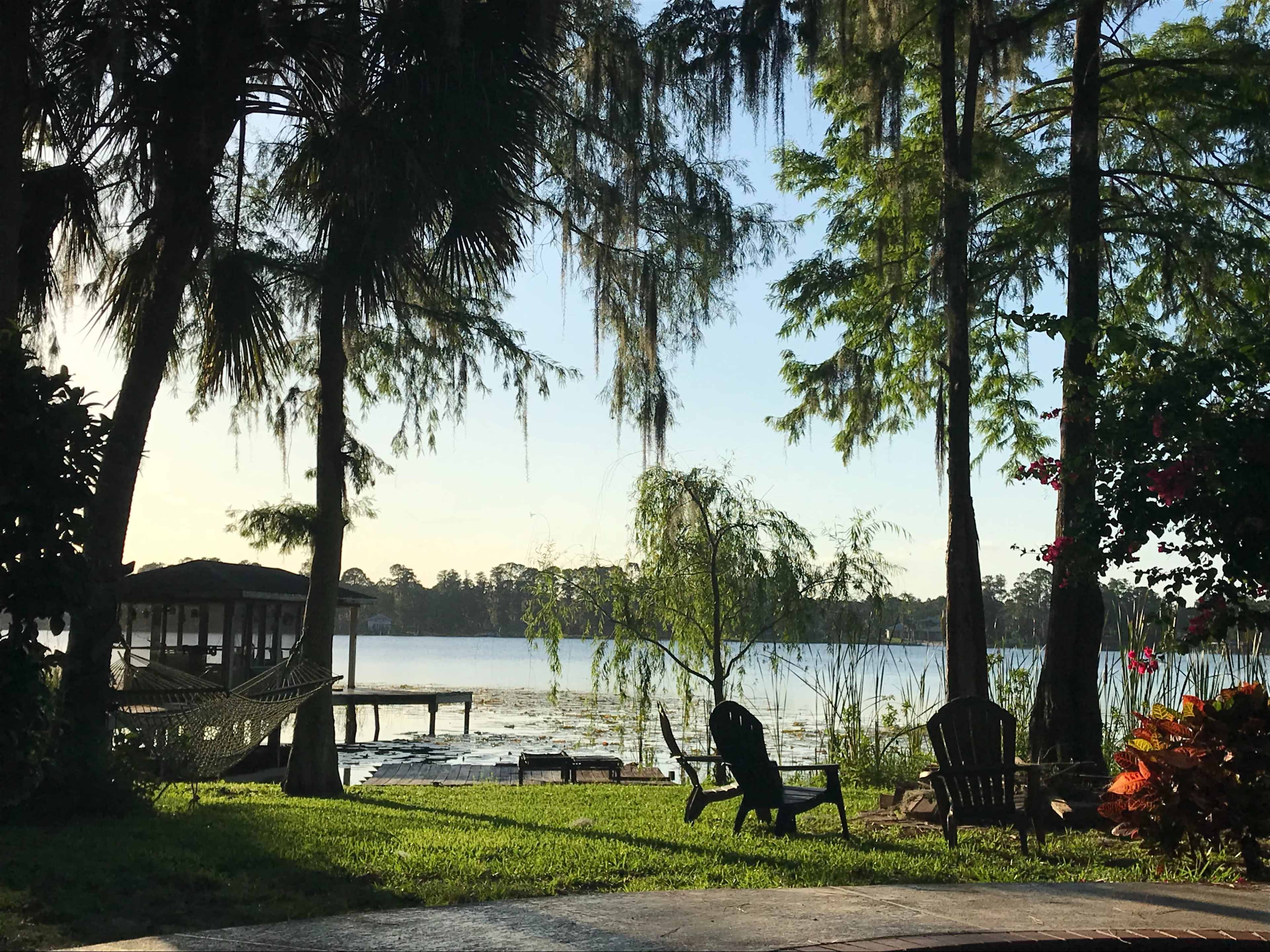 A tranquil lakeside scene with a dock, hammocks, and chairs surrounded by trees.