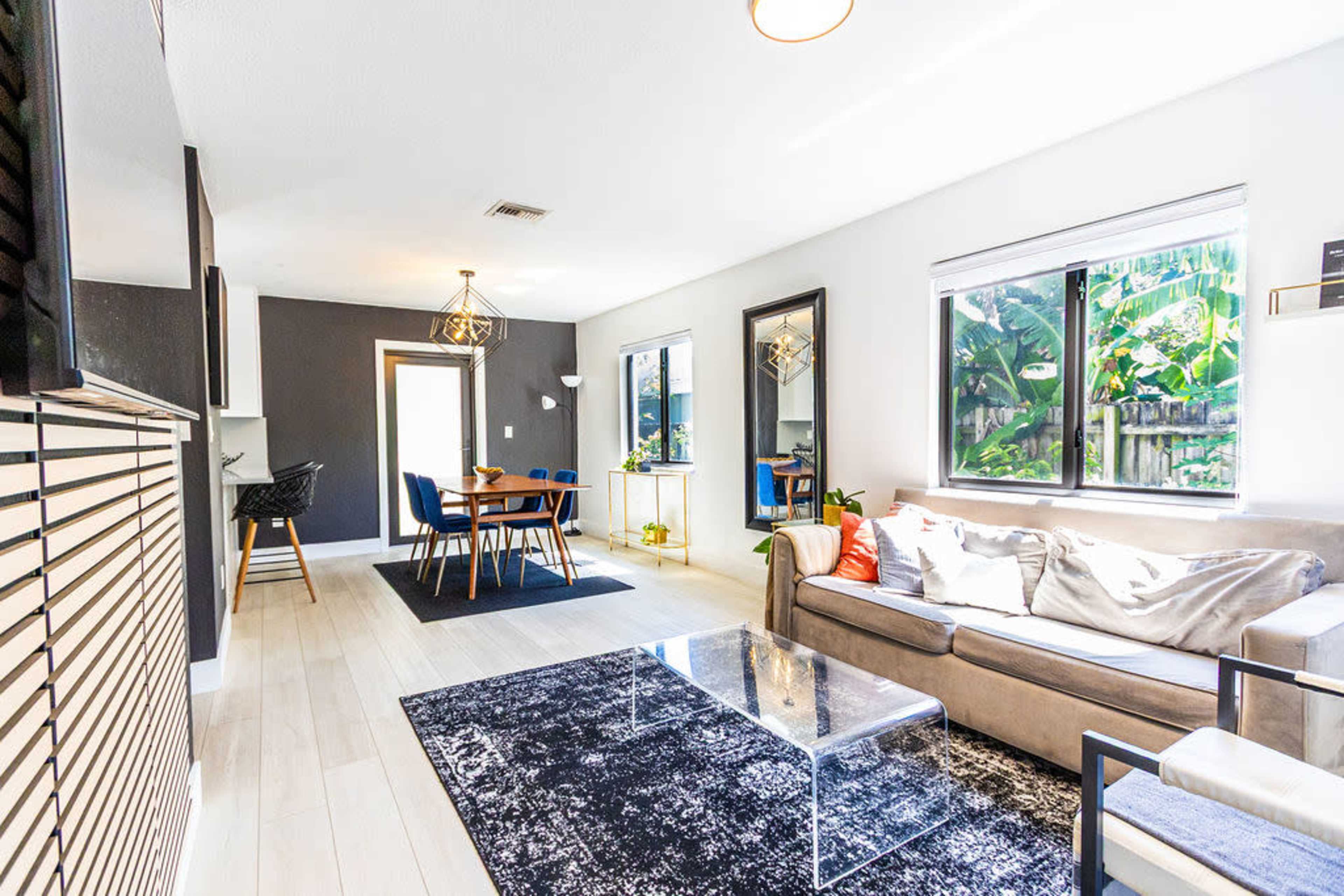 The living room features a modern sofa, a glass coffee table, and a dining area with a wooden table surrounded by chairs, illuminated by natural light from large windows.