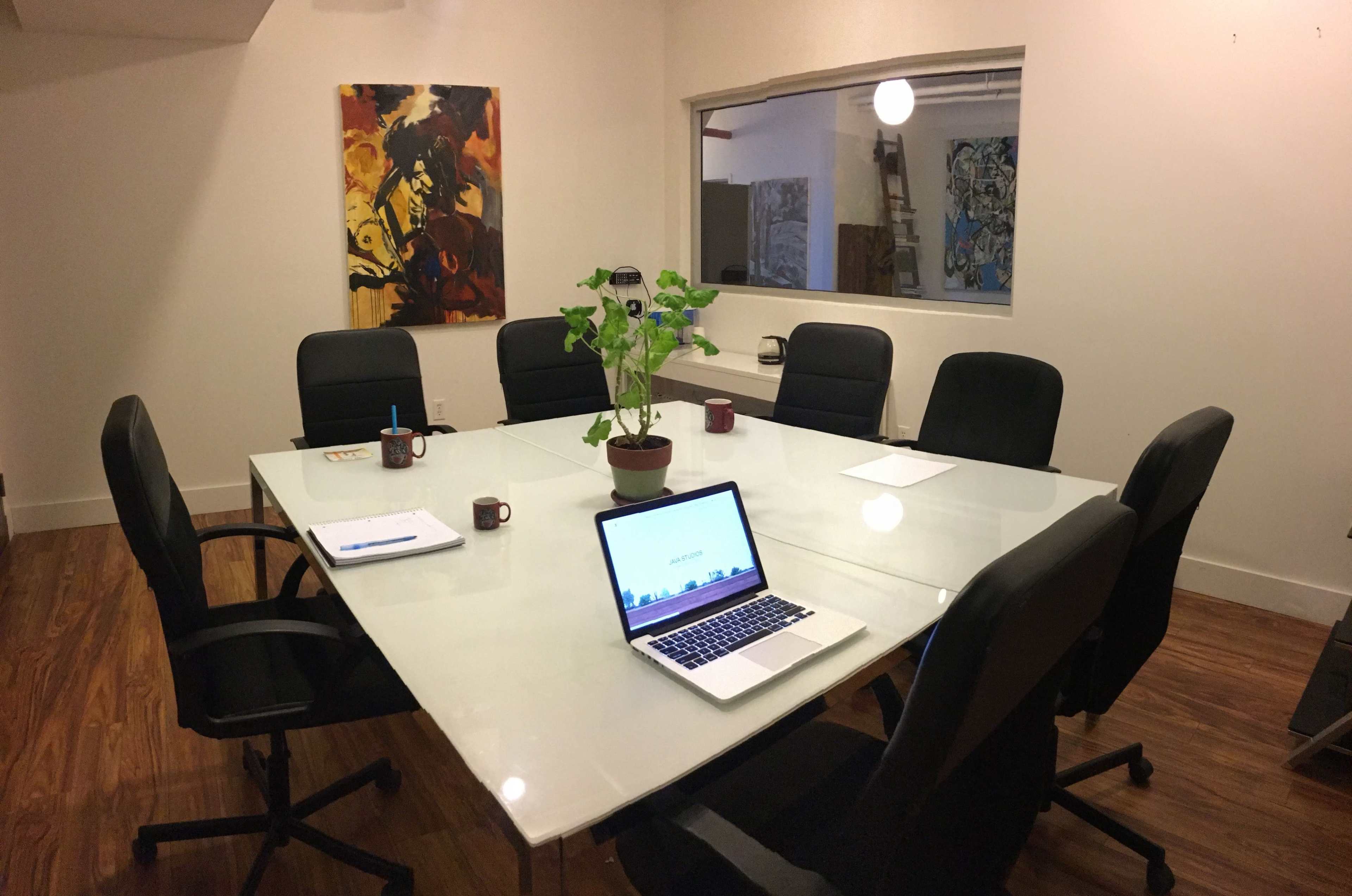 A conference room features a glass table surrounded by black chairs, with a laptop opened on the table and artwork on the walls.