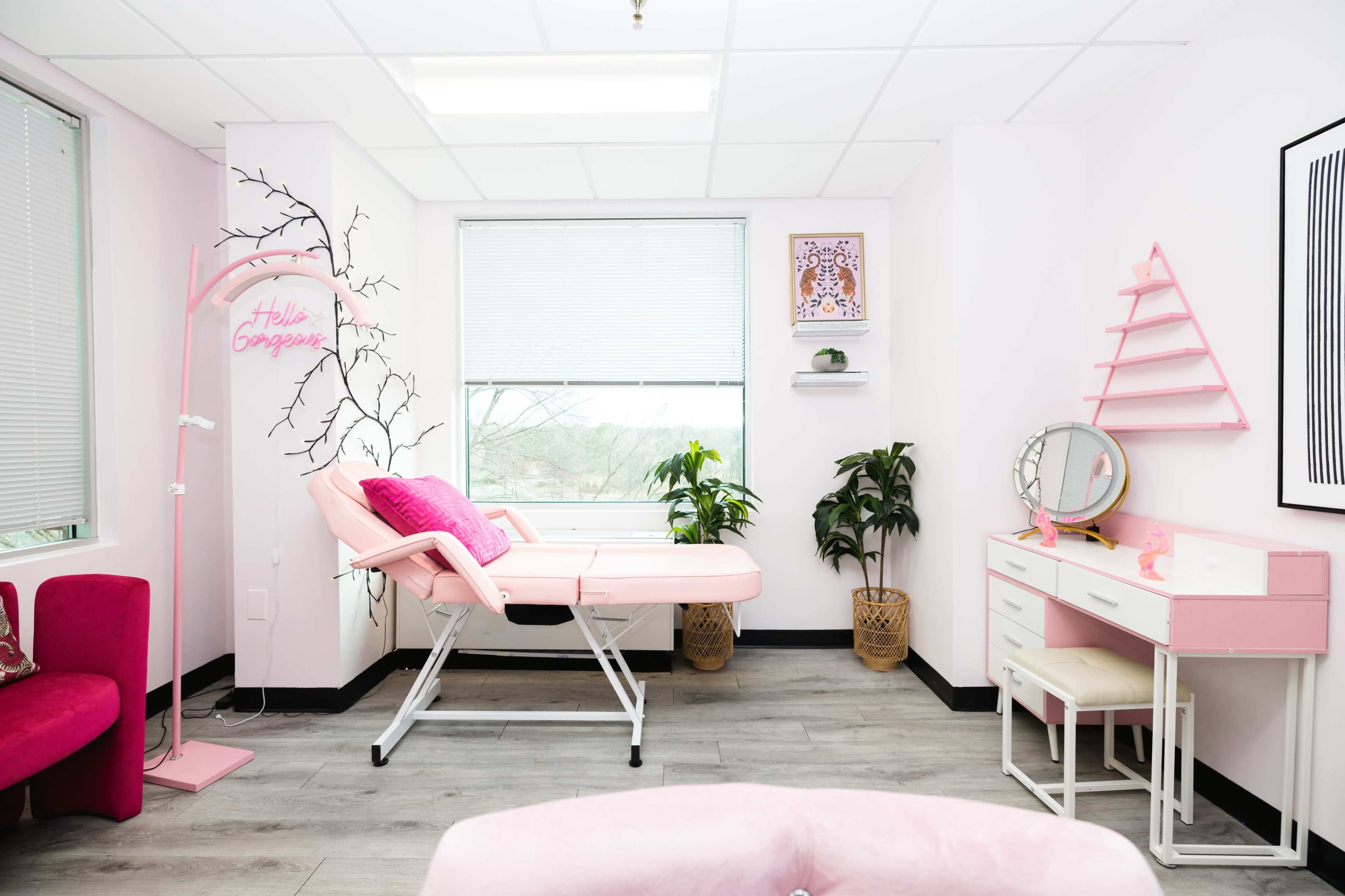 A pink-themed beauty studio with a treatment bed, a vanity mirror, plants, and decorative wall art.