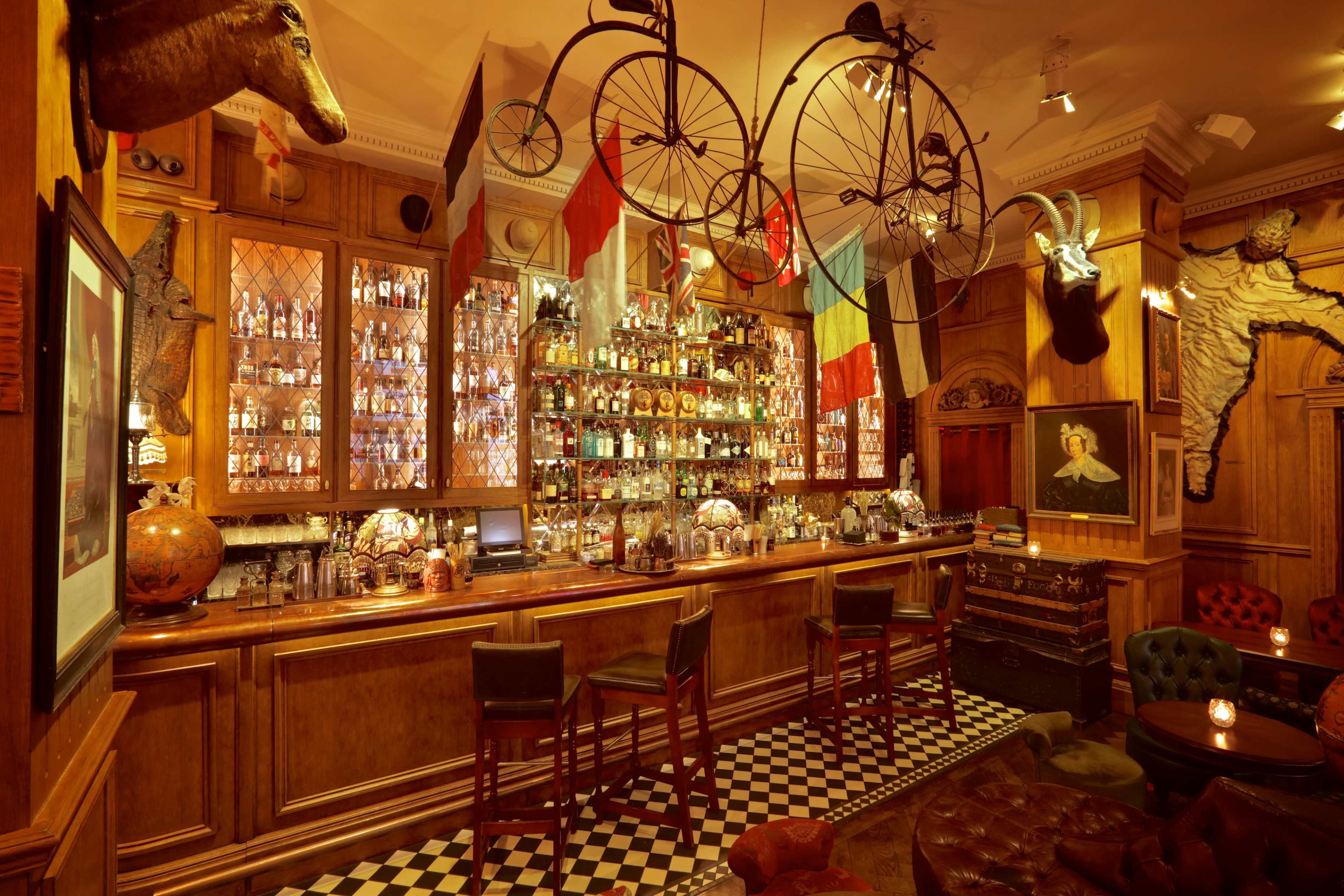 The image shows a vintage-style bar with wooden cabinetry, a variety of bottles displayed behind the counter, and decorative elements like antique bicycles and animal heads on the walls.