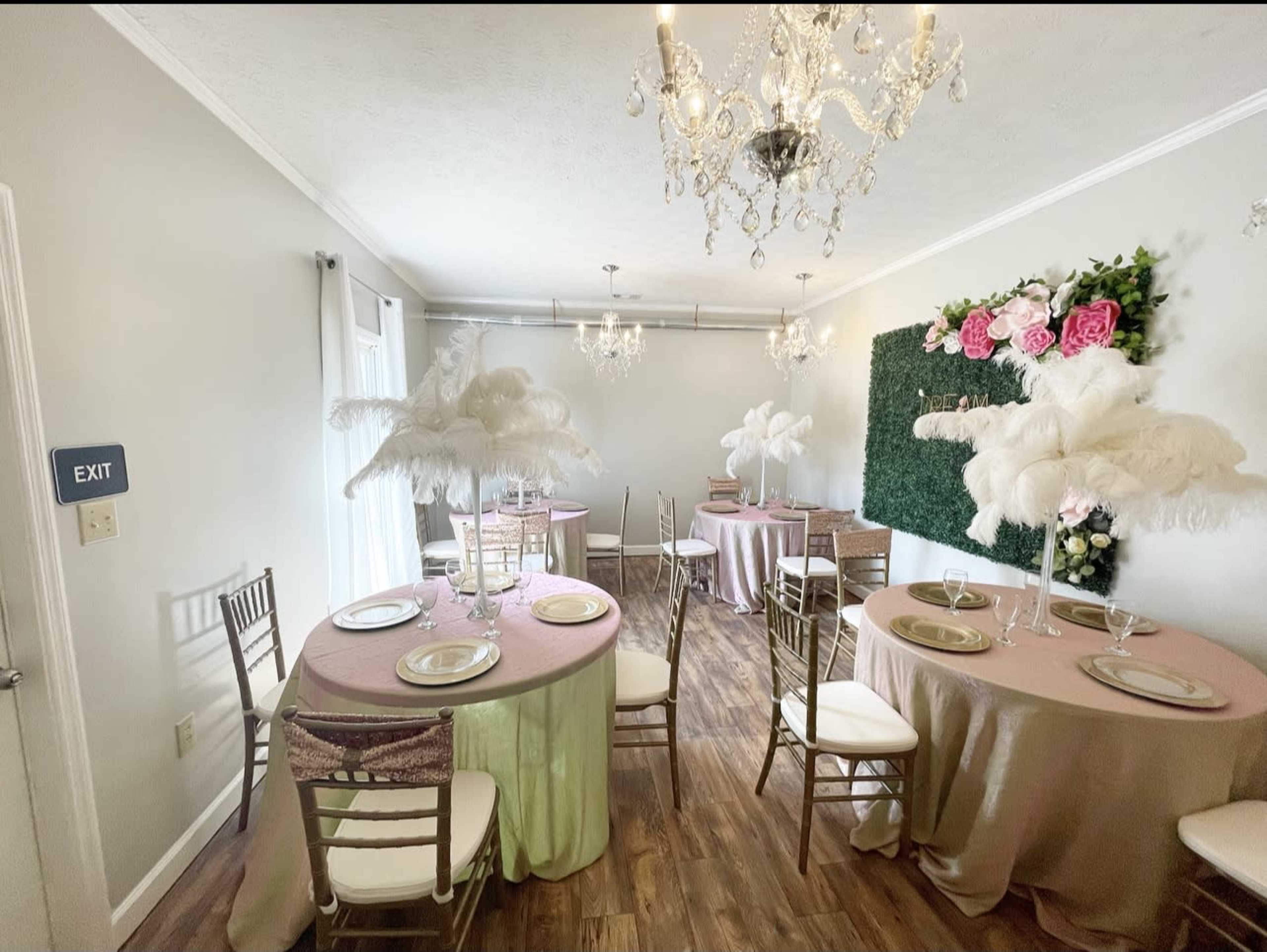 The room features elegantly set tables with pastel tablecloths, adorned with tall feather centerpieces and decorative floral arrangements, under chandeliers.