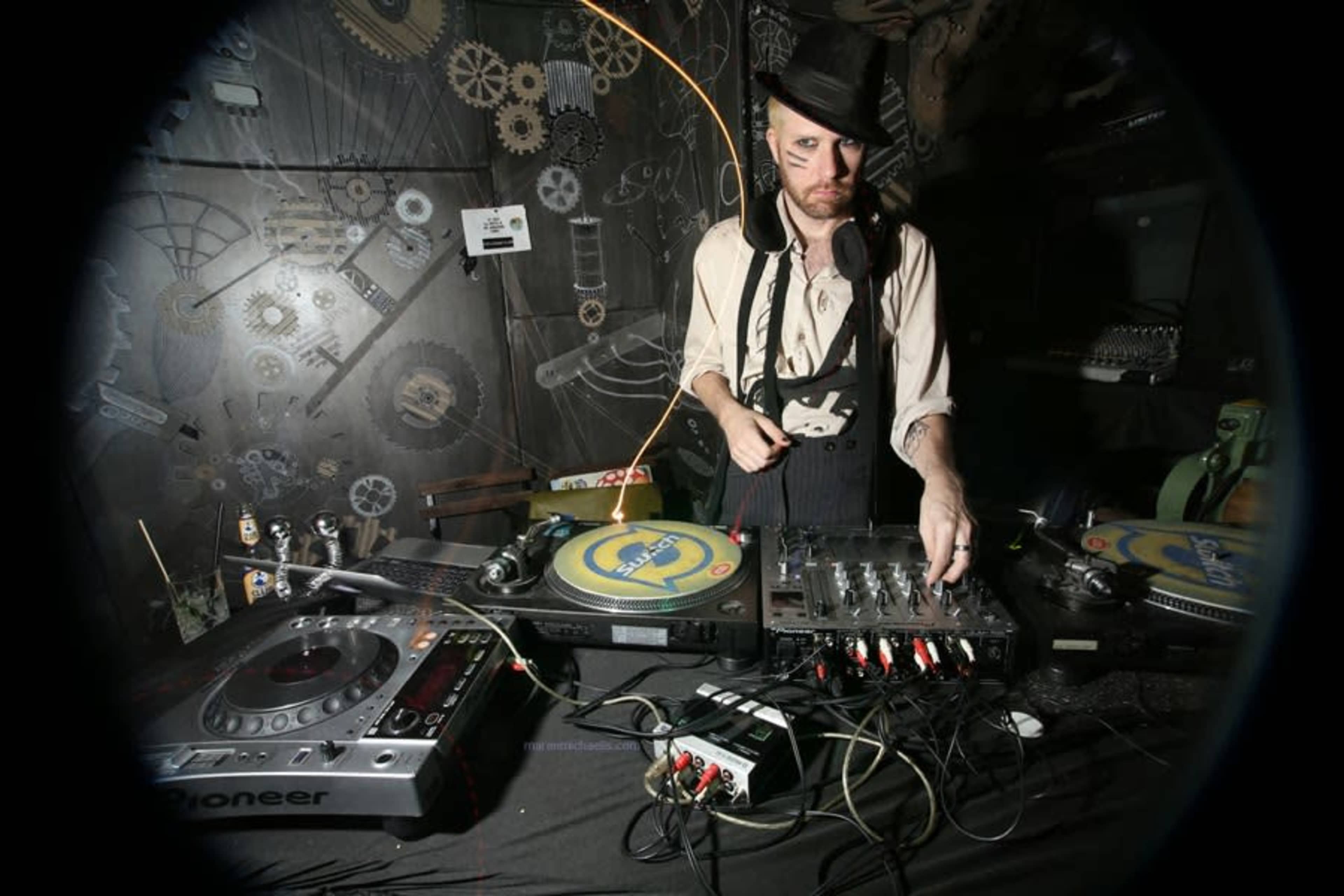 A DJ wearing a hat and makeup operates turntables and a mixer in a dimly lit, industrial-themed setting.