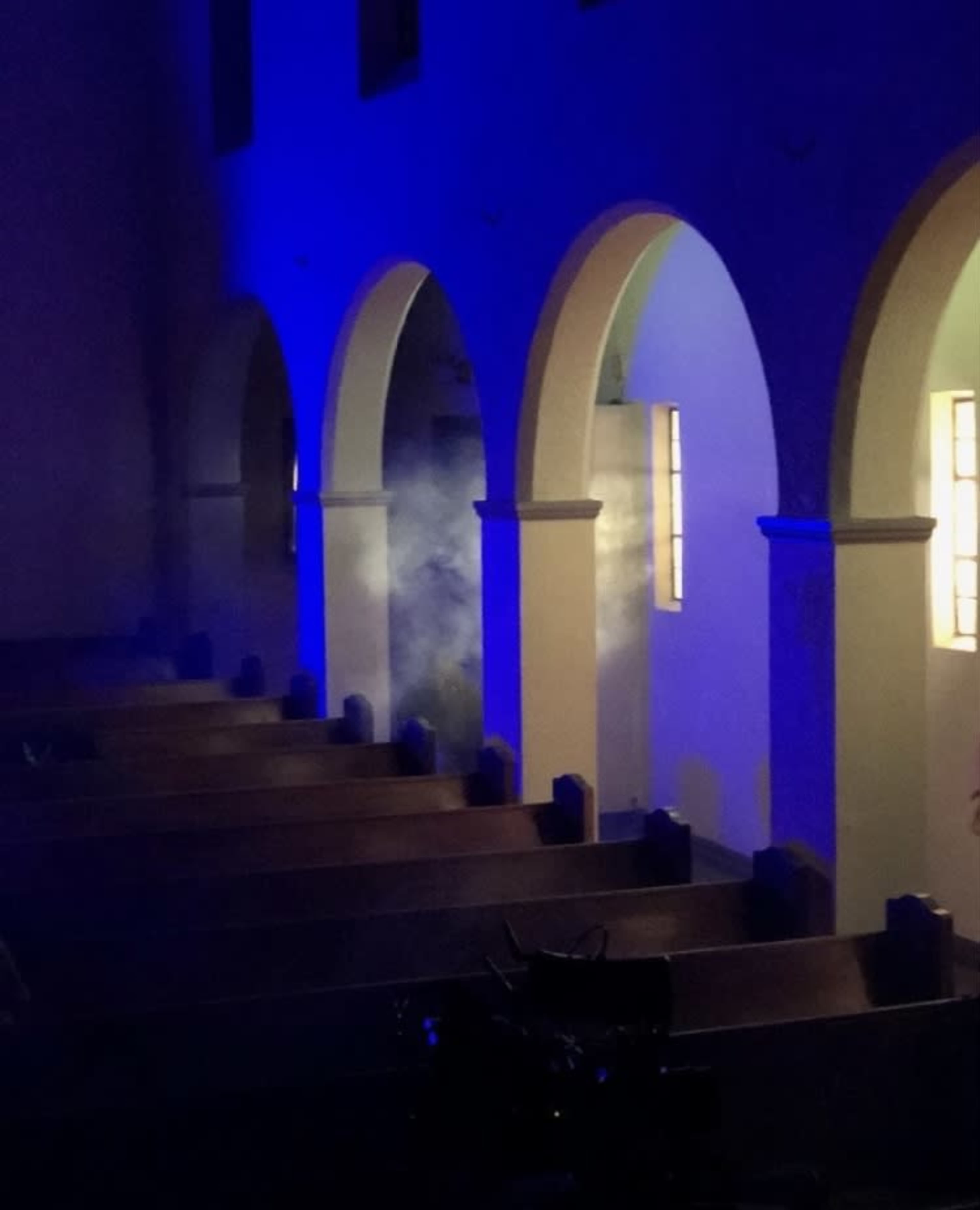 A dimly lit church interior features arched doorways and pews, with blue lighting creating a misty atmosphere.