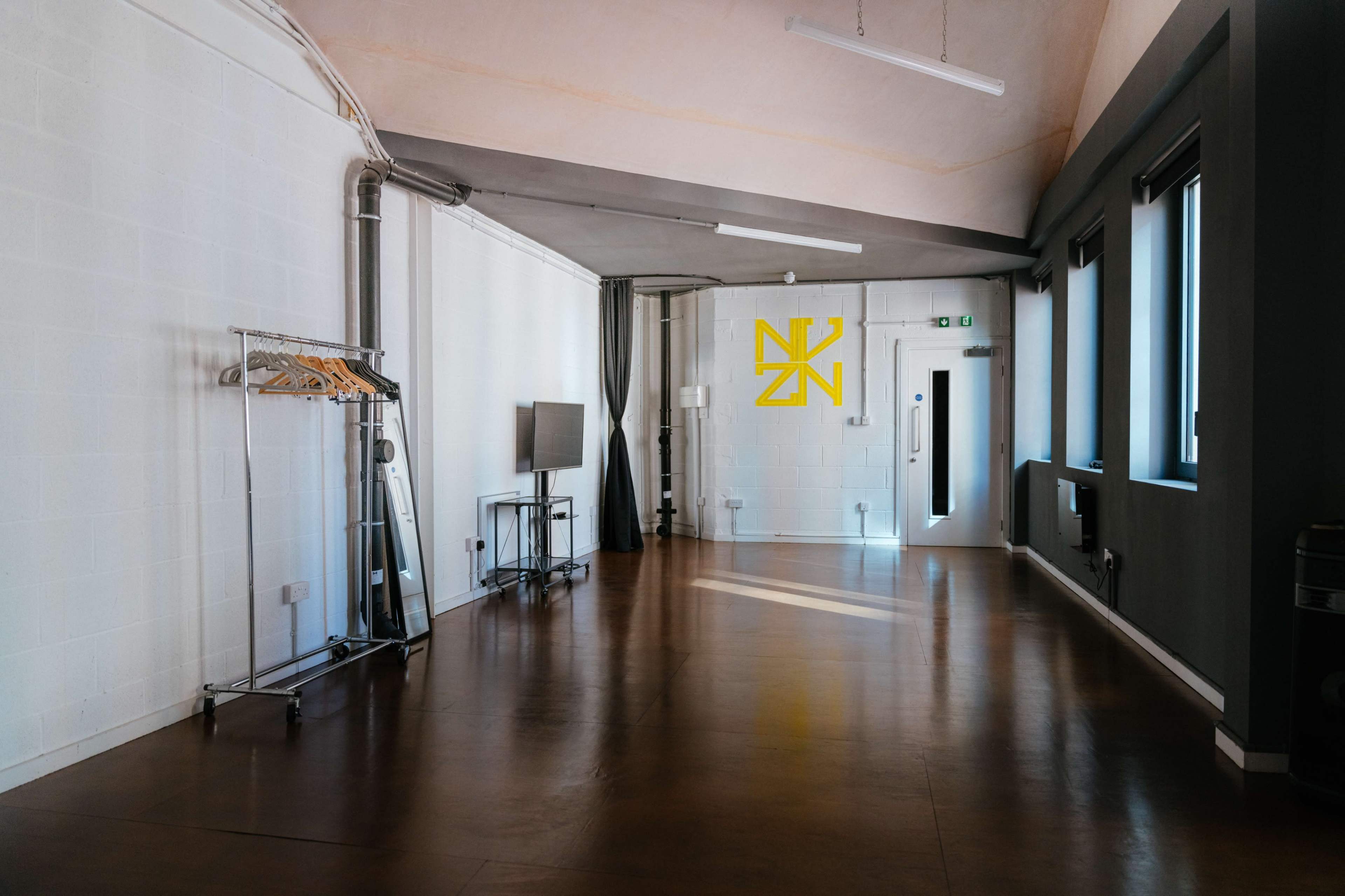 An empty room with a polished floor, a clothing rack on wheels, a television on a stand, and a yellow logo on a white wall.