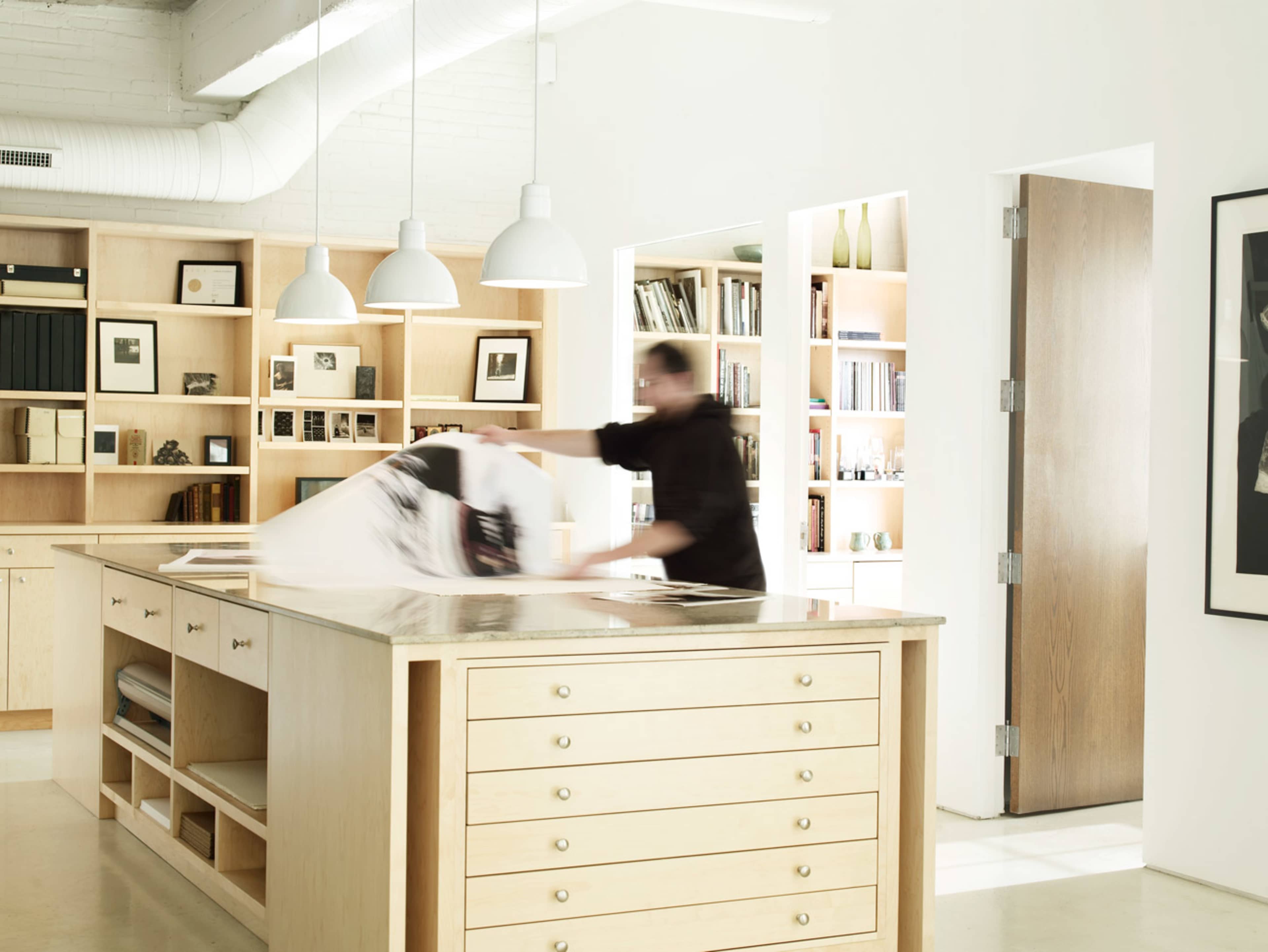 A person is pulling a large piece of fabric off a table in a brightly lit room with wooden shelves and natural wood cabinetry.