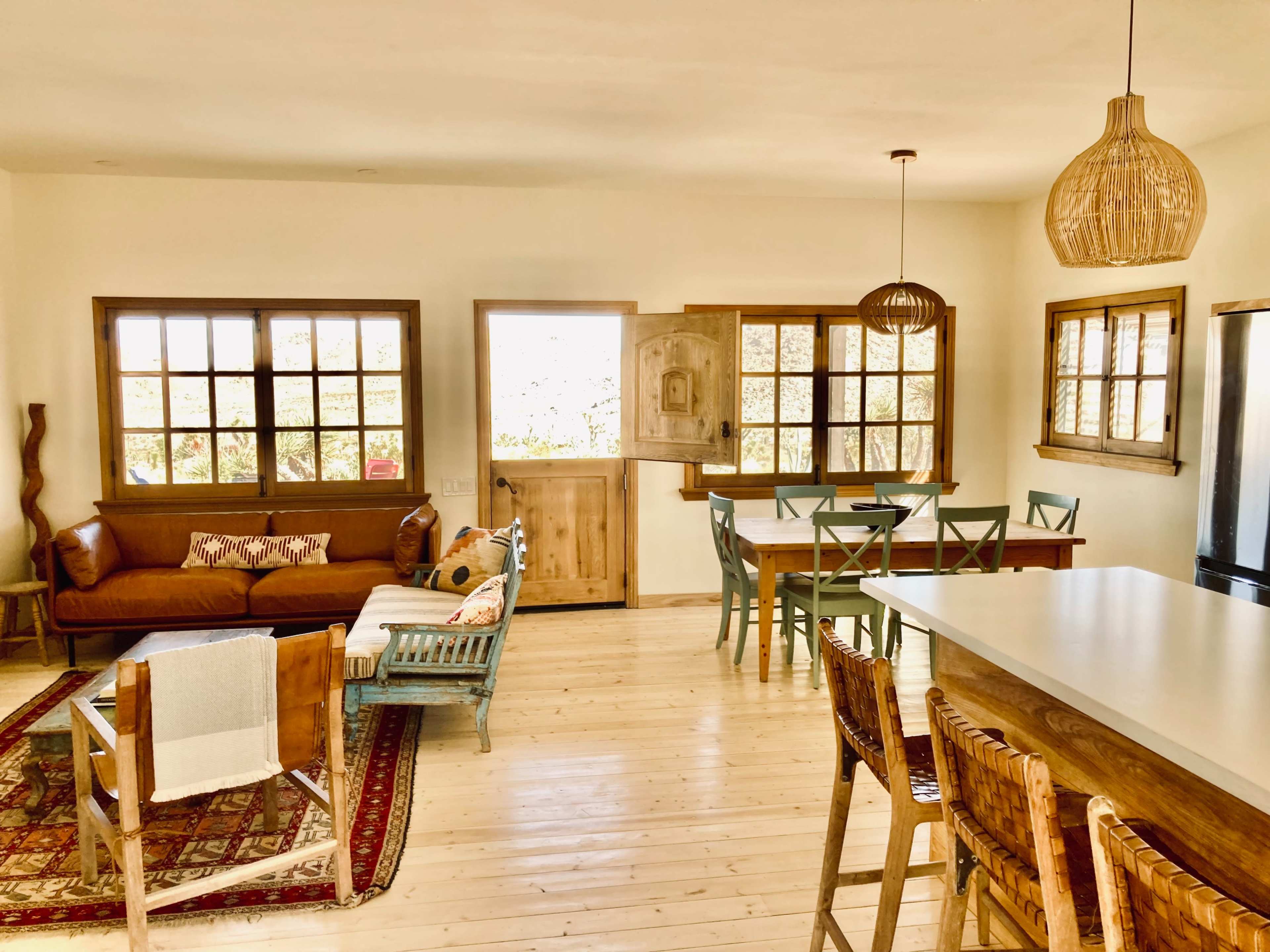 The image shows a bright, open-plan living area with a sofa, dining table, and kitchen bar, featuring wooden furniture and large windows.