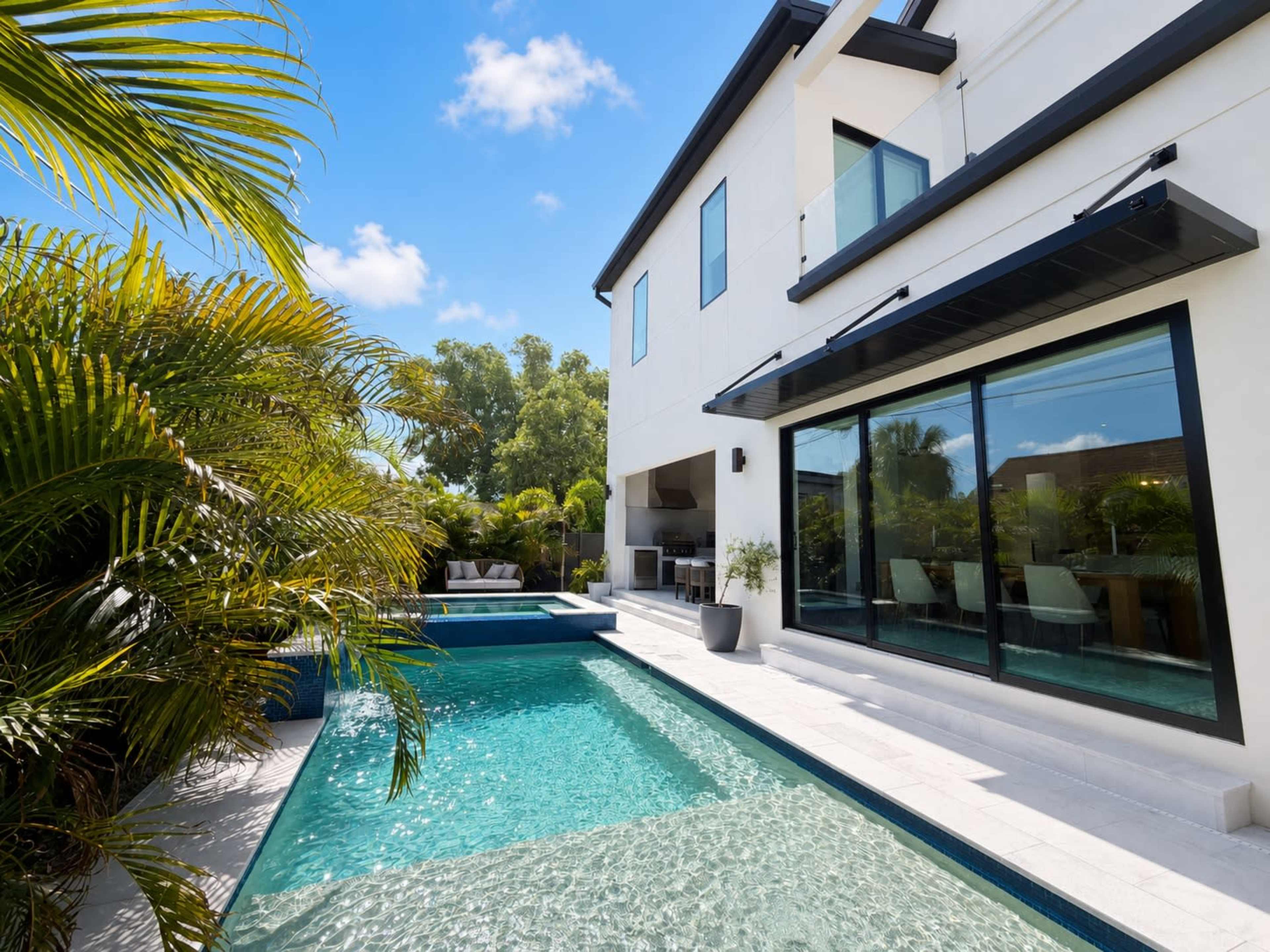 The image shows a modern house with large glass windows beside a swimming pool surrounded by tropical plants.